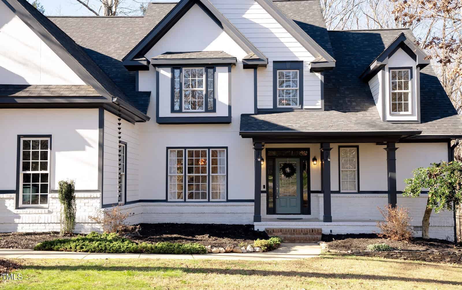 A modern two-story house with white siding, black trim, and multiple large windows. The front porch has columns and a dark door with a wreath. Sparse landscaping and leafless trees suggest late autumn or winter.