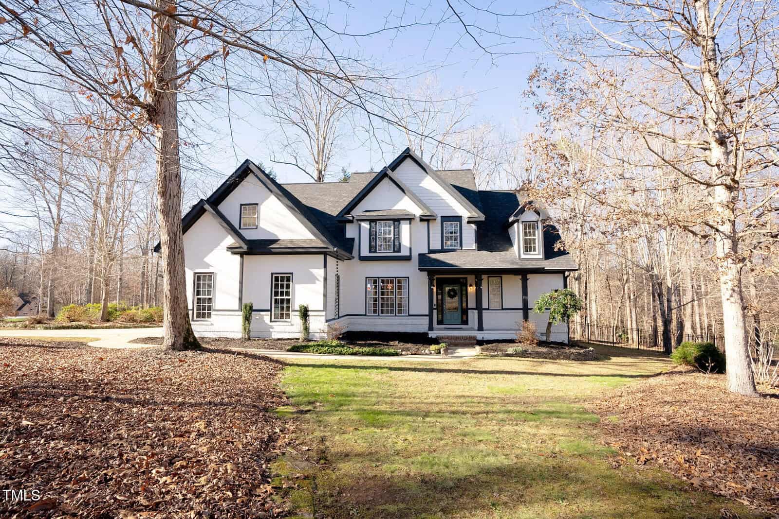 A two-story white house with black trim and a front porch sits among leafless trees on a sunny day. A driveway curves to the left, and the lawn is covered with brown leaves.