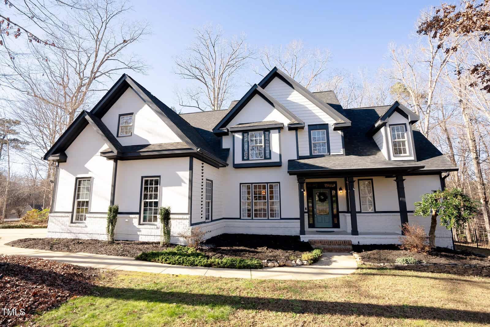 A two-story white house with black trim, multiple gables, and a covered front porch, surrounded by bare trees and a neatly landscaped yard under a clear sky.