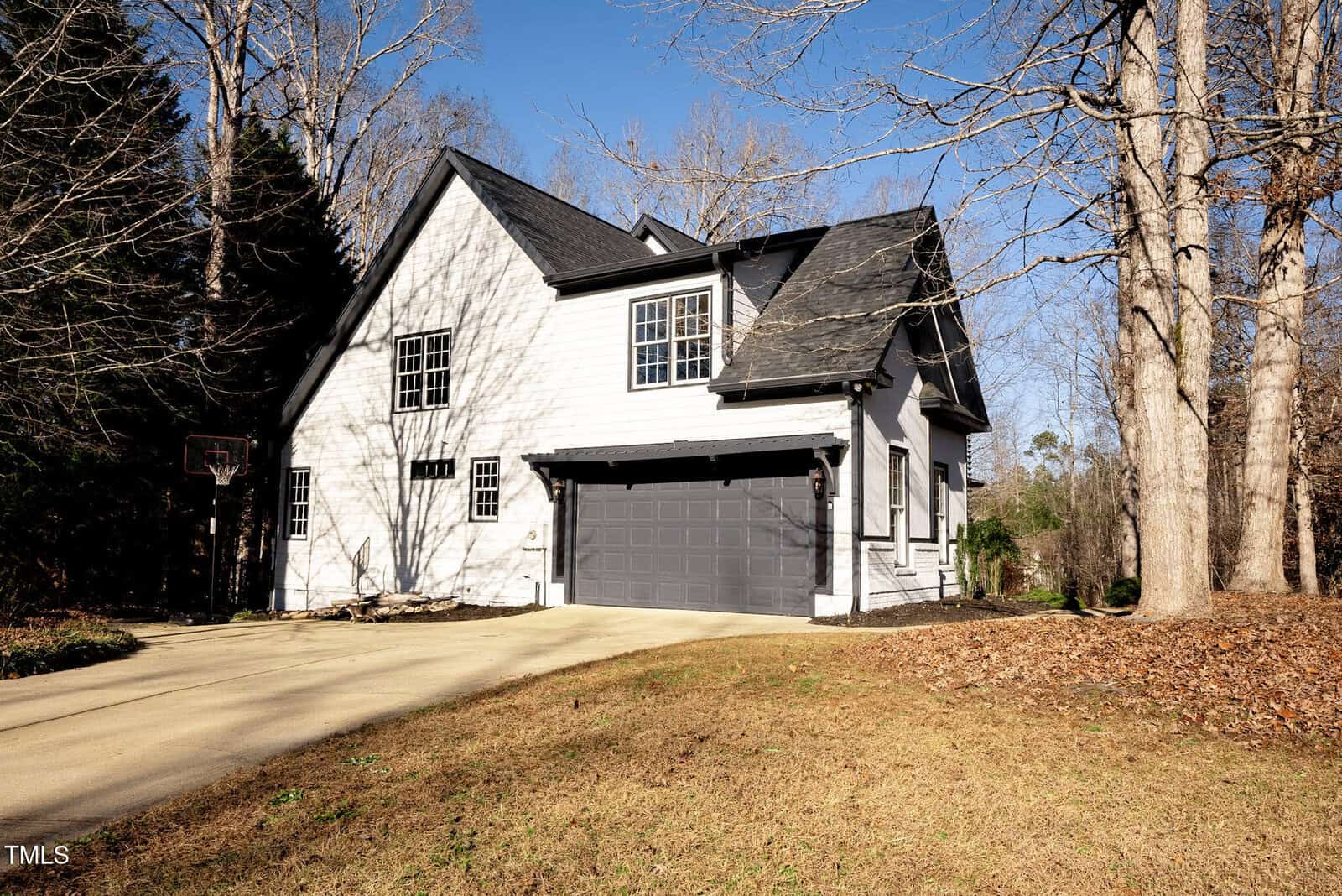 A two-story white house with black trim and a dark gray double garage sits on a sloped driveway, surrounded by bare trees and a grassy yard under a clear blue sky.