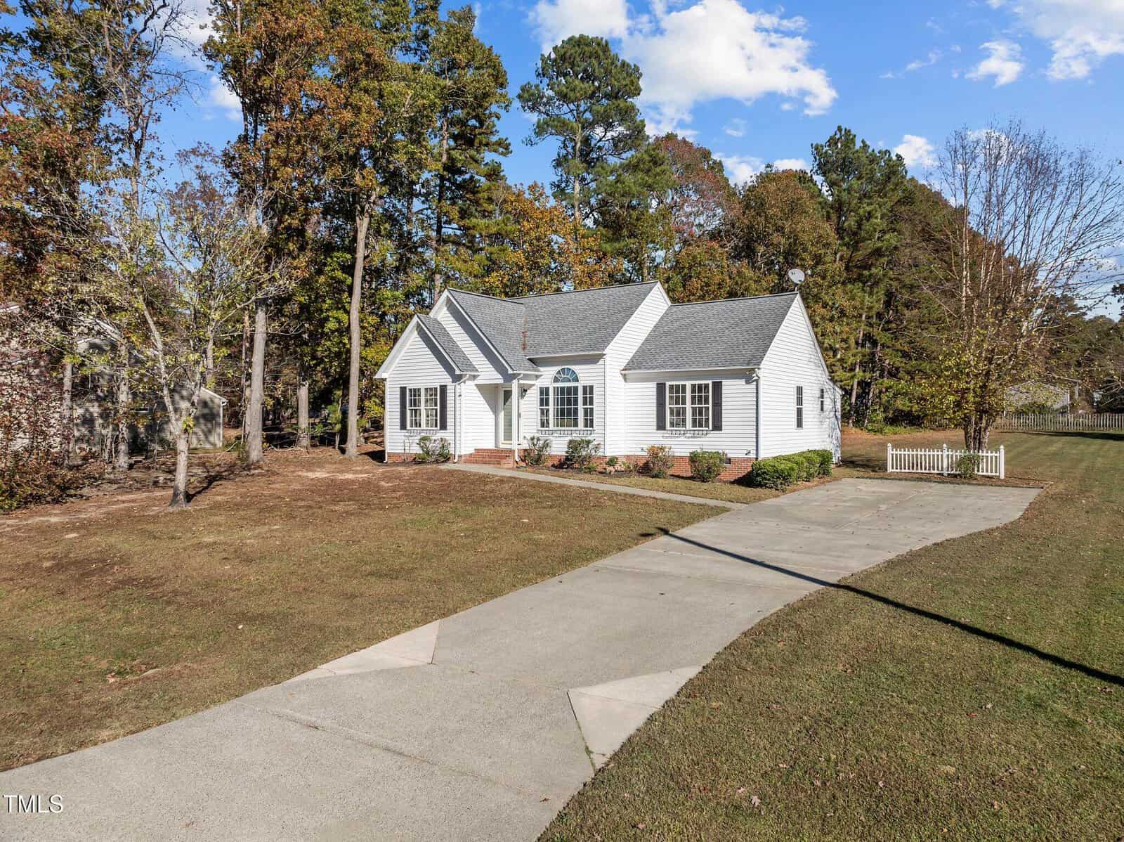 A single-story white house with gray roof shingles sits at the end of a long concrete driveway, surrounded by grass, trees with autumn foliage, and a white picket fence on the right side.
