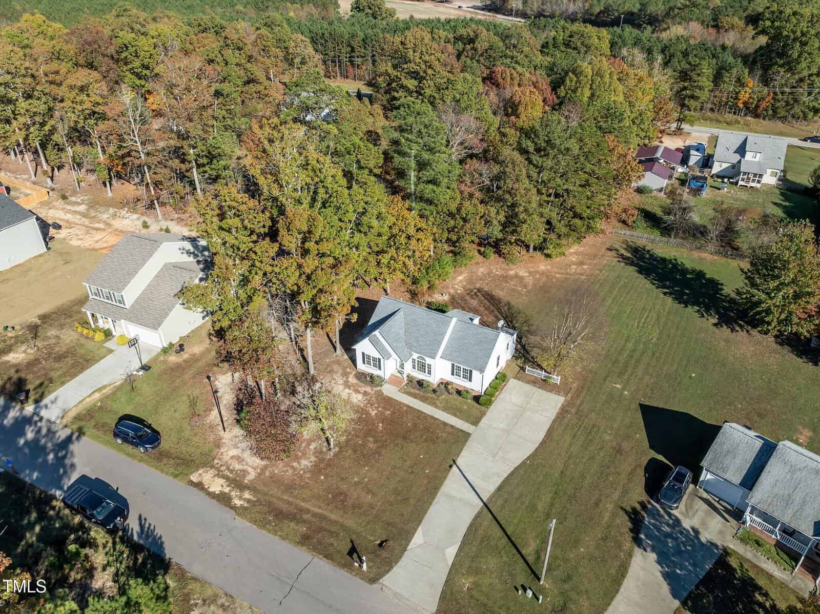 Aerial view of a single-story white house with a gray roof, a driveway, and a large grassy yard. The property is surrounded by trees and neighboring homes on a suburban street.