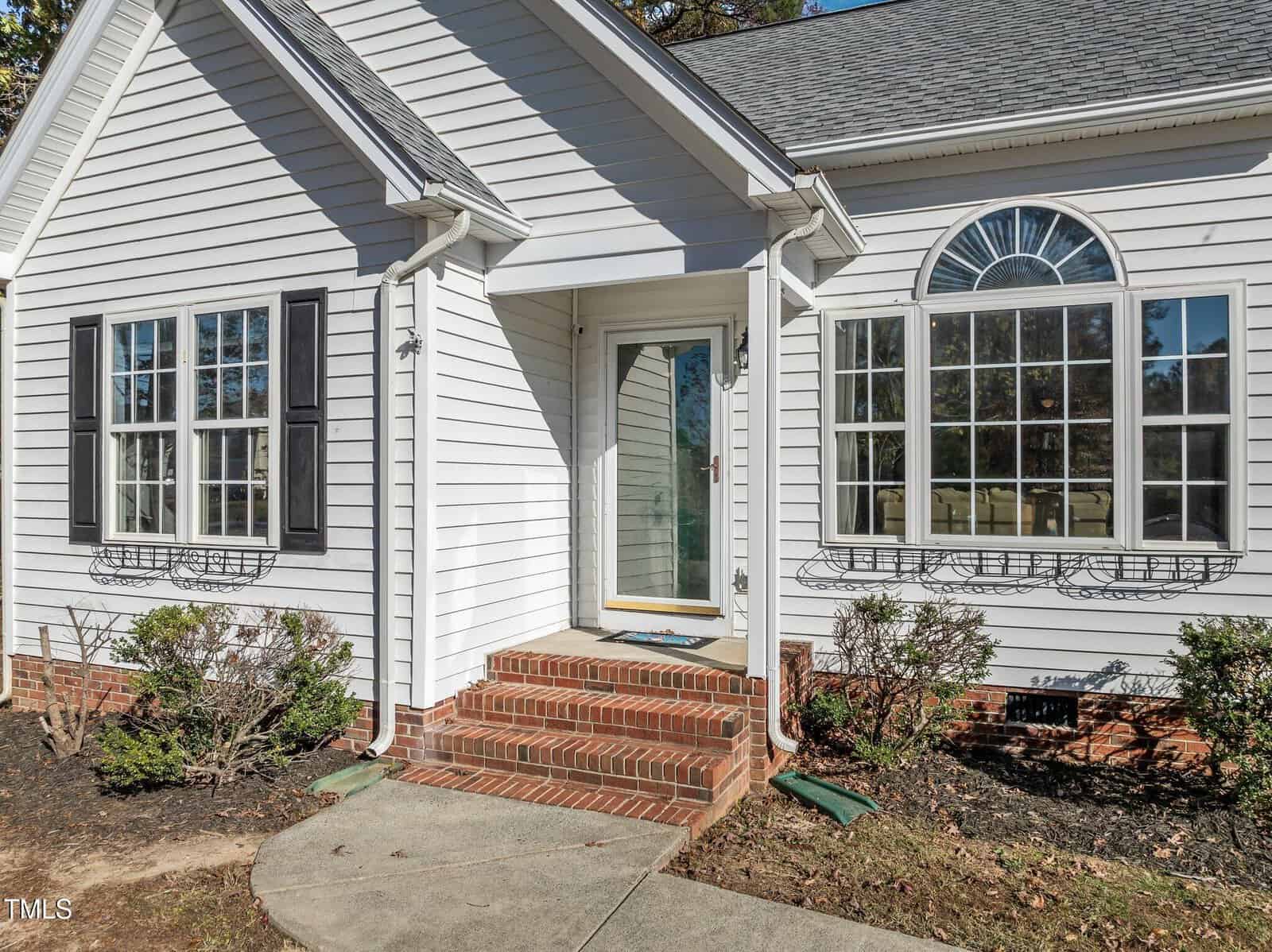 A white house with brick steps leading to a glass front door, black shutters on the windows, and some small shrubs planted along the foundation. The yard has a paved walkway and mulched areas.