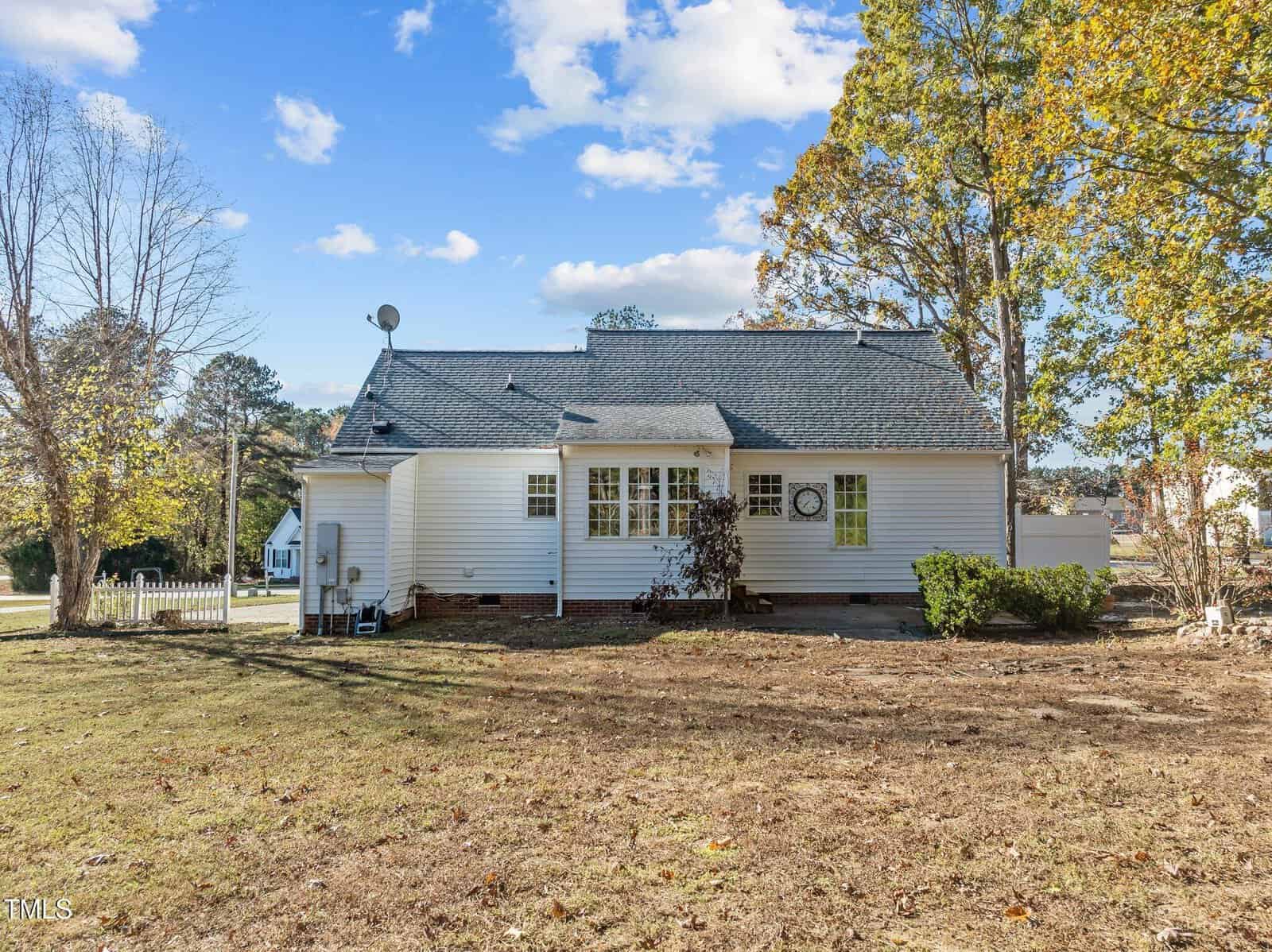 White house with a dark roof, several windows, and a satellite dish. The yard is mostly bare with some grass and trees, and the sky is blue with a few clouds.