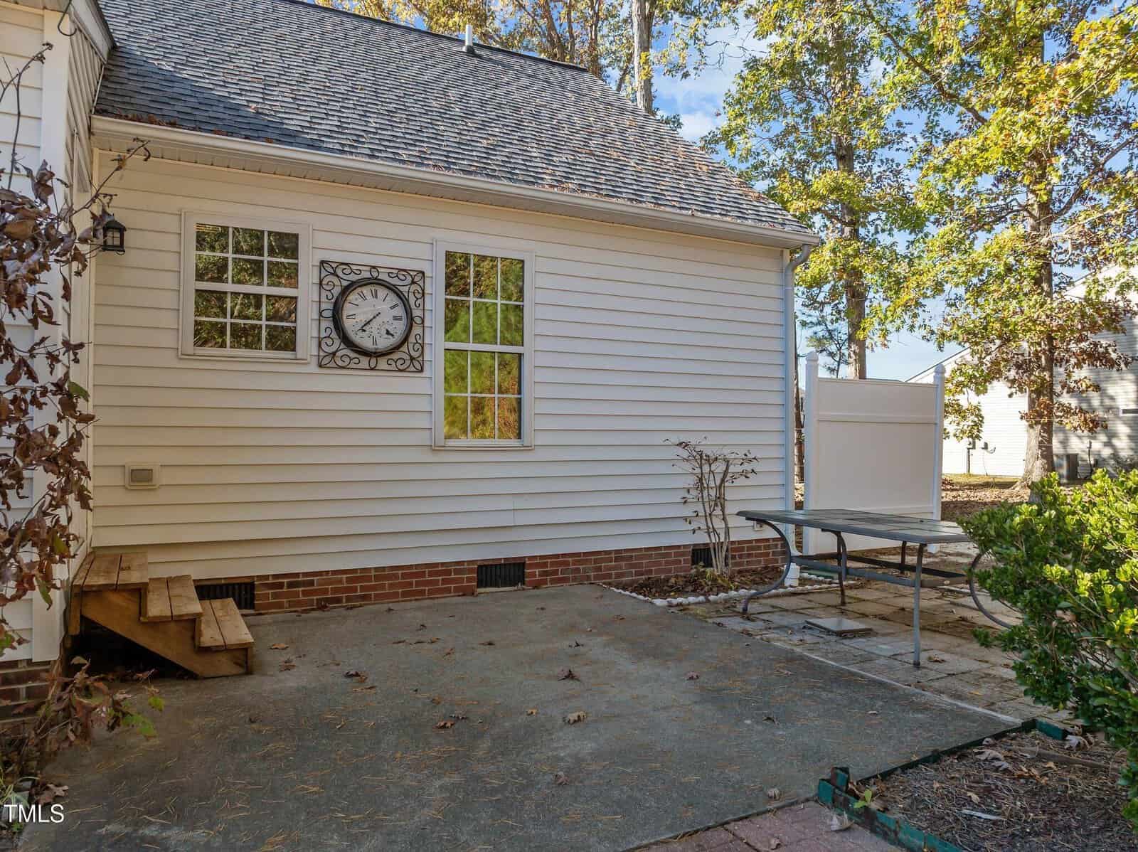 A small backyard patio with a metal table and bench, bare shrubs, and steps leading to a white-sided house. A large decorative clock hangs between two windows. Trees with autumn leaves are visible in the background.