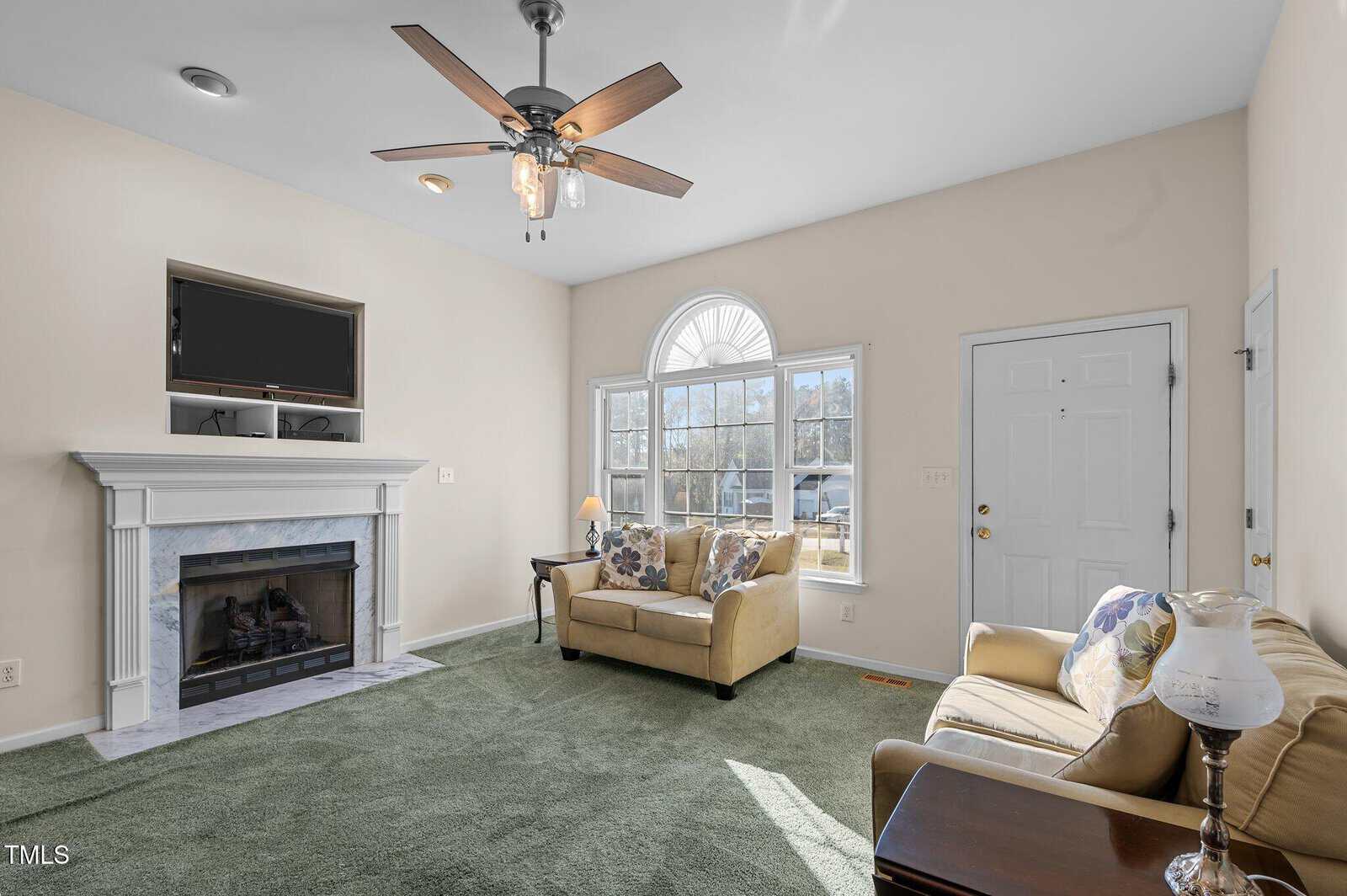 Living room with green carpet, beige walls, a white fireplace, a TV above the mantle, two beige sofas near a large arched window, a ceiling fan, and a white front door.