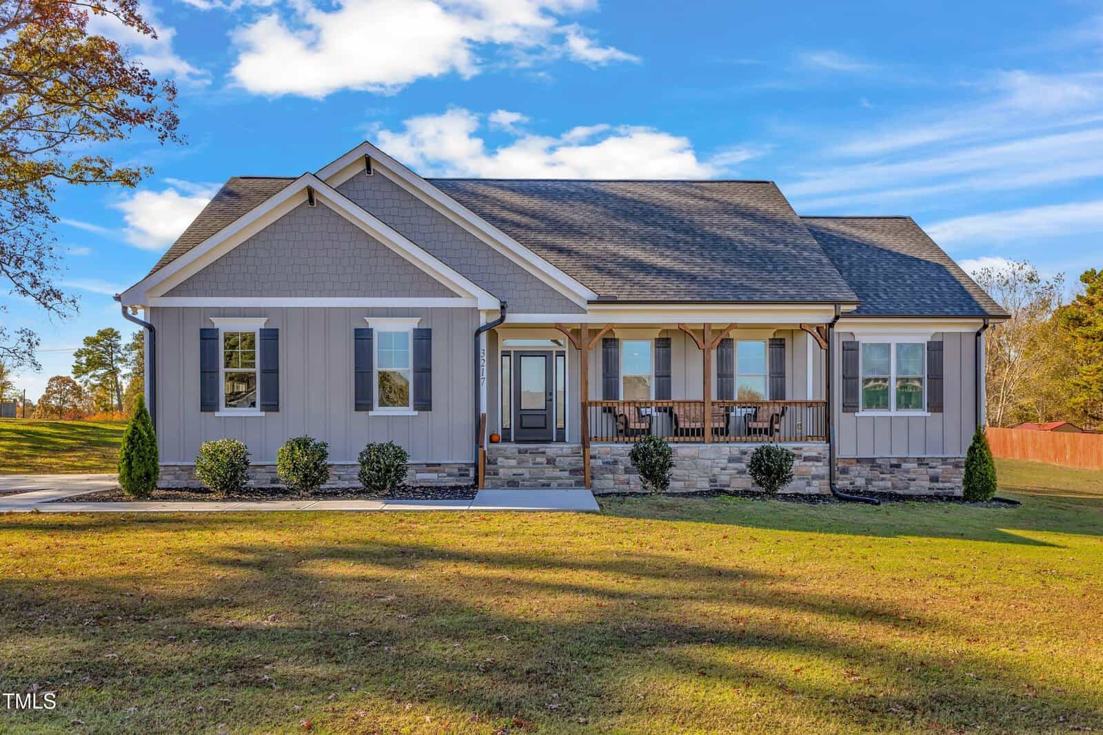 A single-story grey house with a dark roof sits under a blue sky. The front features a porch with wooden columns and a few chairs. A path leads to the entrance, surrounded by a neatly trimmed lawn and small shrubs.