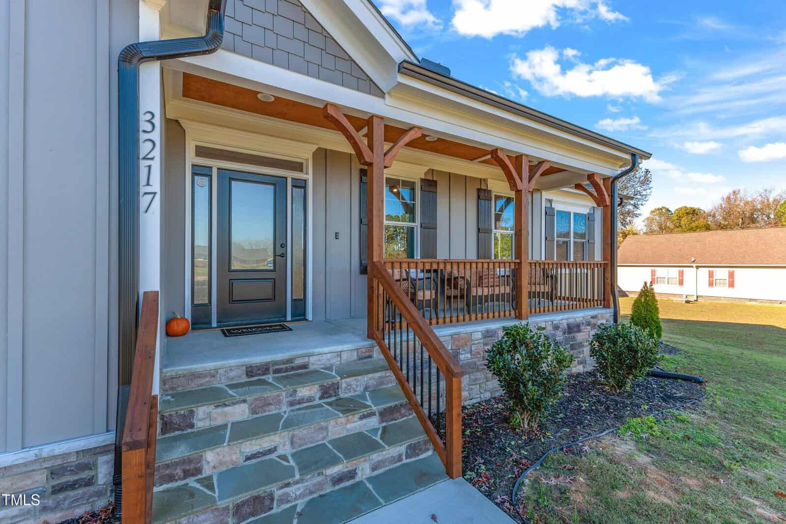 A modern house with a stone facade and wooden accents. The porch features a welcoming entrance with a glass door, a pumpkin decoration, and a small seating area. Blue skies and a neighboring building are visible in the background.