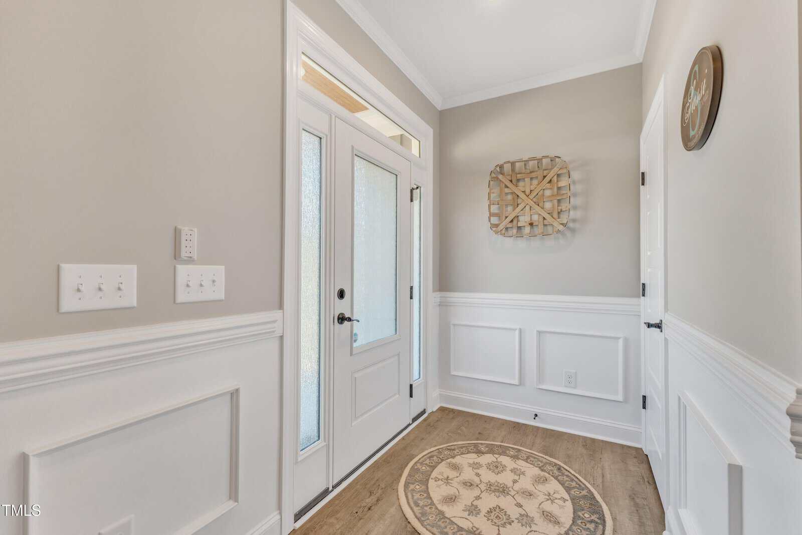 A bright entryway with a glass-paneled door, white wainscoting, and beige walls. A round rug with a floral pattern is on the floor. A decorative wall piece and a clock are mounted on the walls.