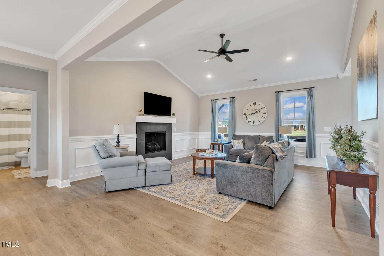 A cozy living room with light wood flooring, a gray sectional sofa, and an armchair facing a fireplace with a TV above. A large clock hangs near two windows with sheer curtains, and a small side table features a potted plant.