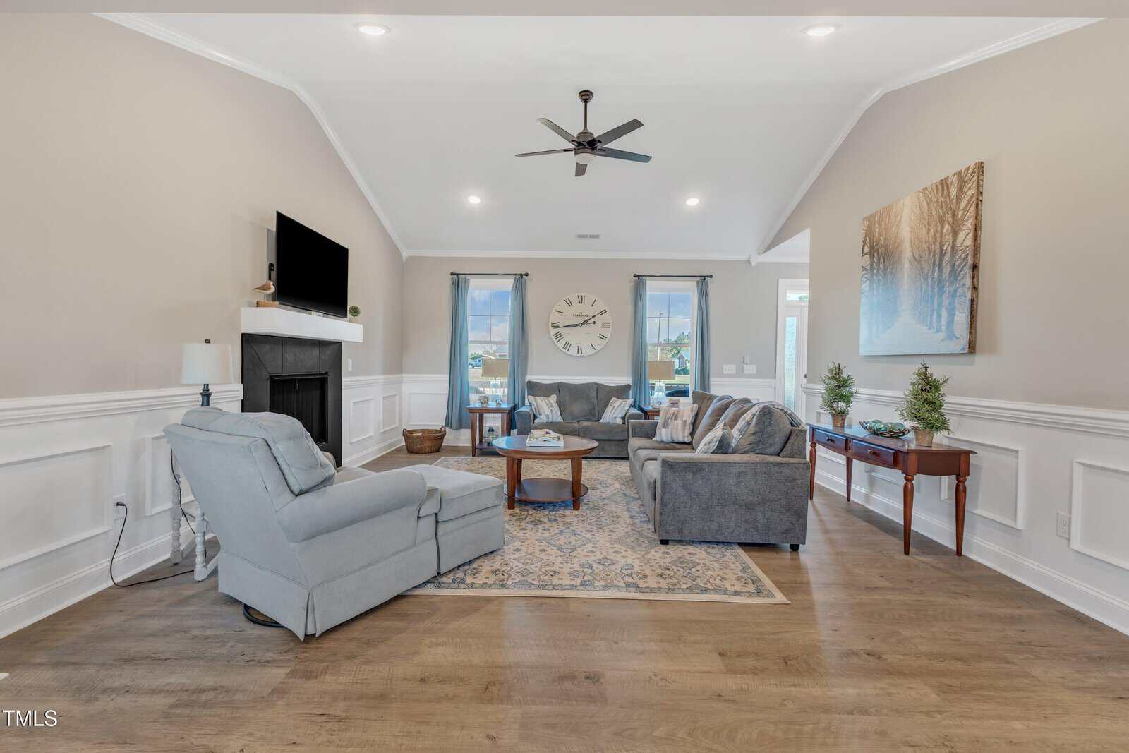 A modern living room with beige walls and wood flooring, featuring a gray sofa, armchairs, a fireplace with a TV above it, and a large wall clock. Two windows with curtains allow natural light to illuminate the space.
