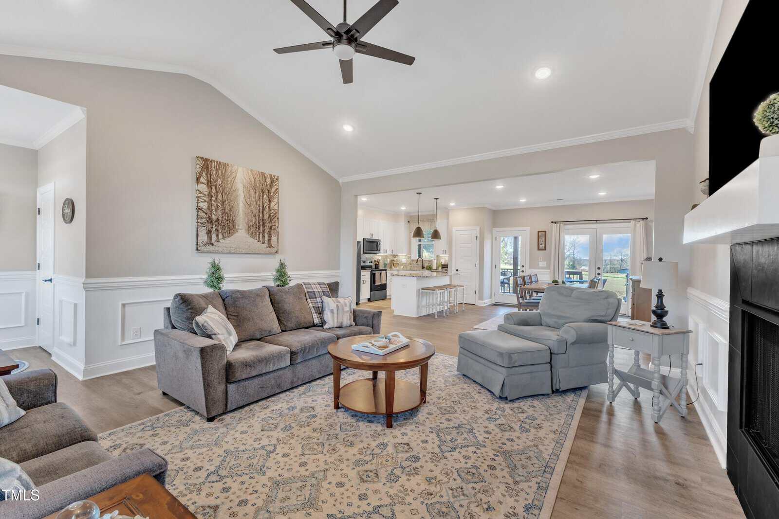 Modern living room with neutral tones, featuring a gray sofa and armchair, a round wooden coffee table on a patterned rug, and a fireplace. The open layout leads to a kitchen with a large island and a dining area with a clear view of the outdoors.
