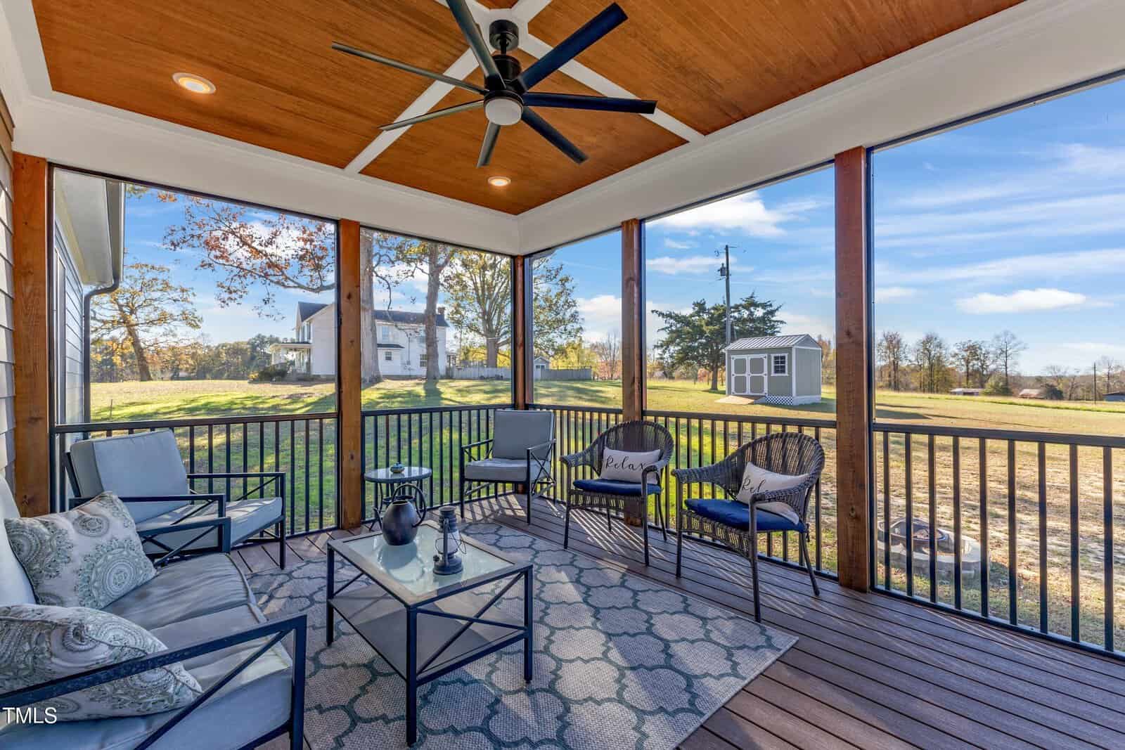 A screened porch with patio furniture including chairs and a table with a teapot and cups. The porch has a wooden ceiling with a fan. The view features a yard, trees, and a distant white house under a blue sky with scattered clouds.