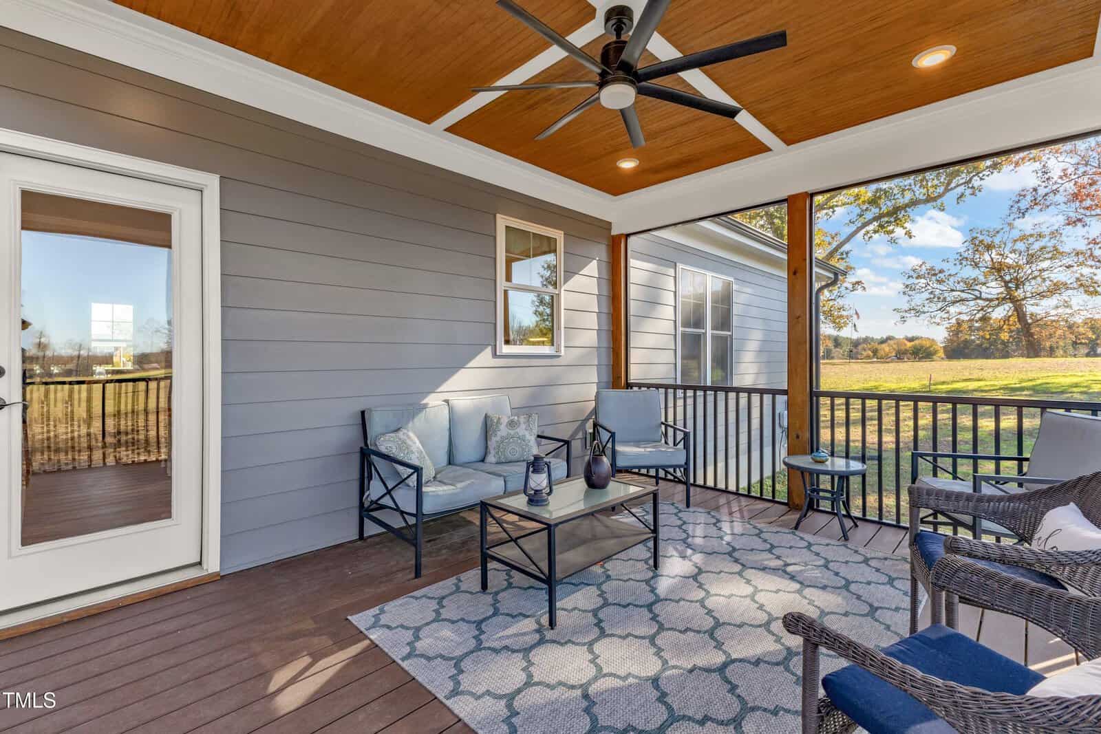 Covered porch with wooden ceiling and fan. Features a seating area with a sofa, chairs, and a small table on a patterned rug. Glass doors and windows provide views of a grassy landscape with trees. Natural light illuminates the space.