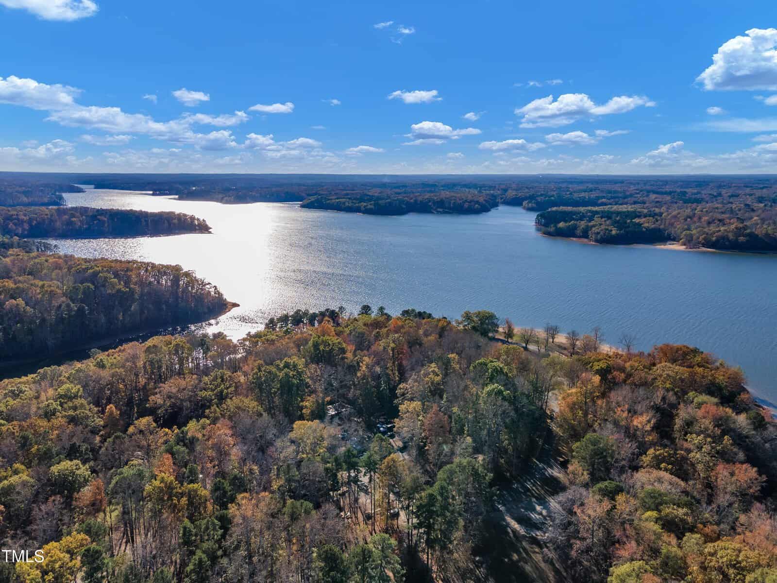 Aerial view of Henderson's lake near 3217 Satterwhite Point Road, surrounded by dense autumn forest. Sunlight reflects off the calm water, forming a bright path across the surface. Trees display a mix of green, orange, and brown foliage beneath a blue sky with scattered clouds.