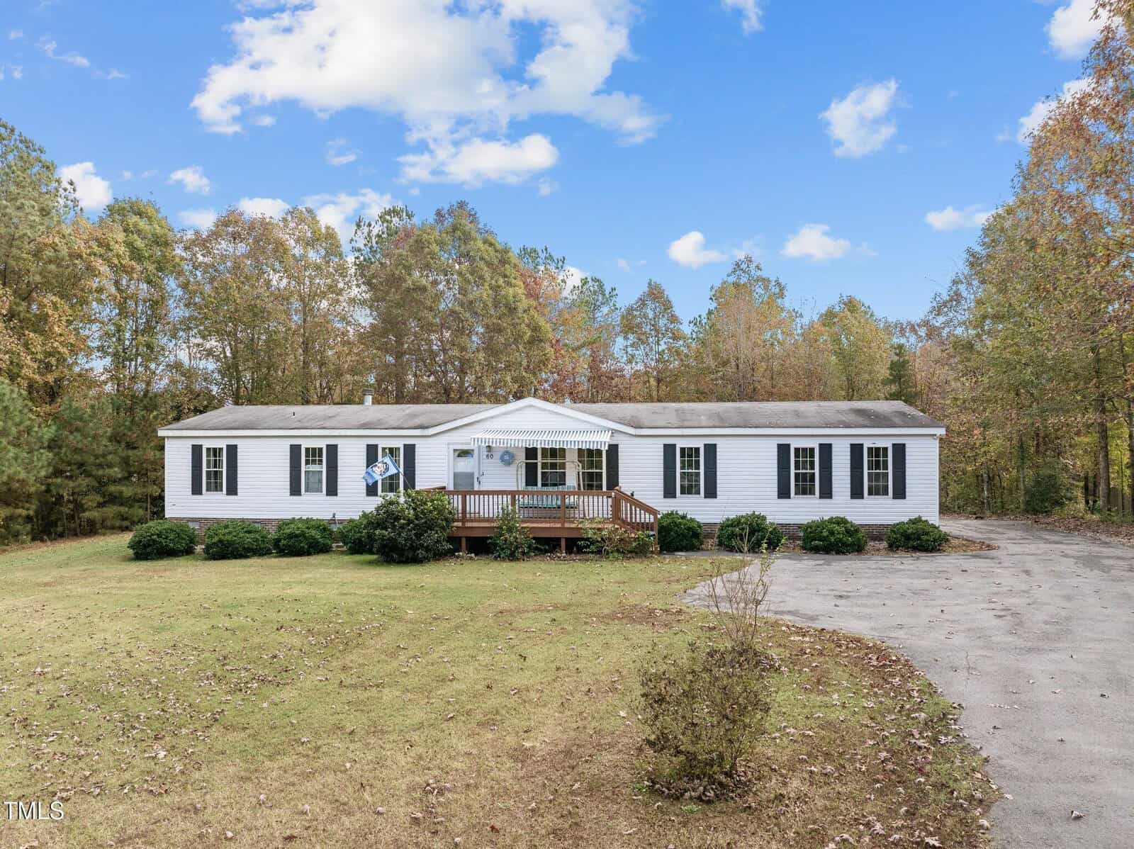 A single-story white manufactured home with a front porch sits on a large grassy lot surrounded by trees. A curved driveway runs along the right side of the house under a partly cloudy sky.