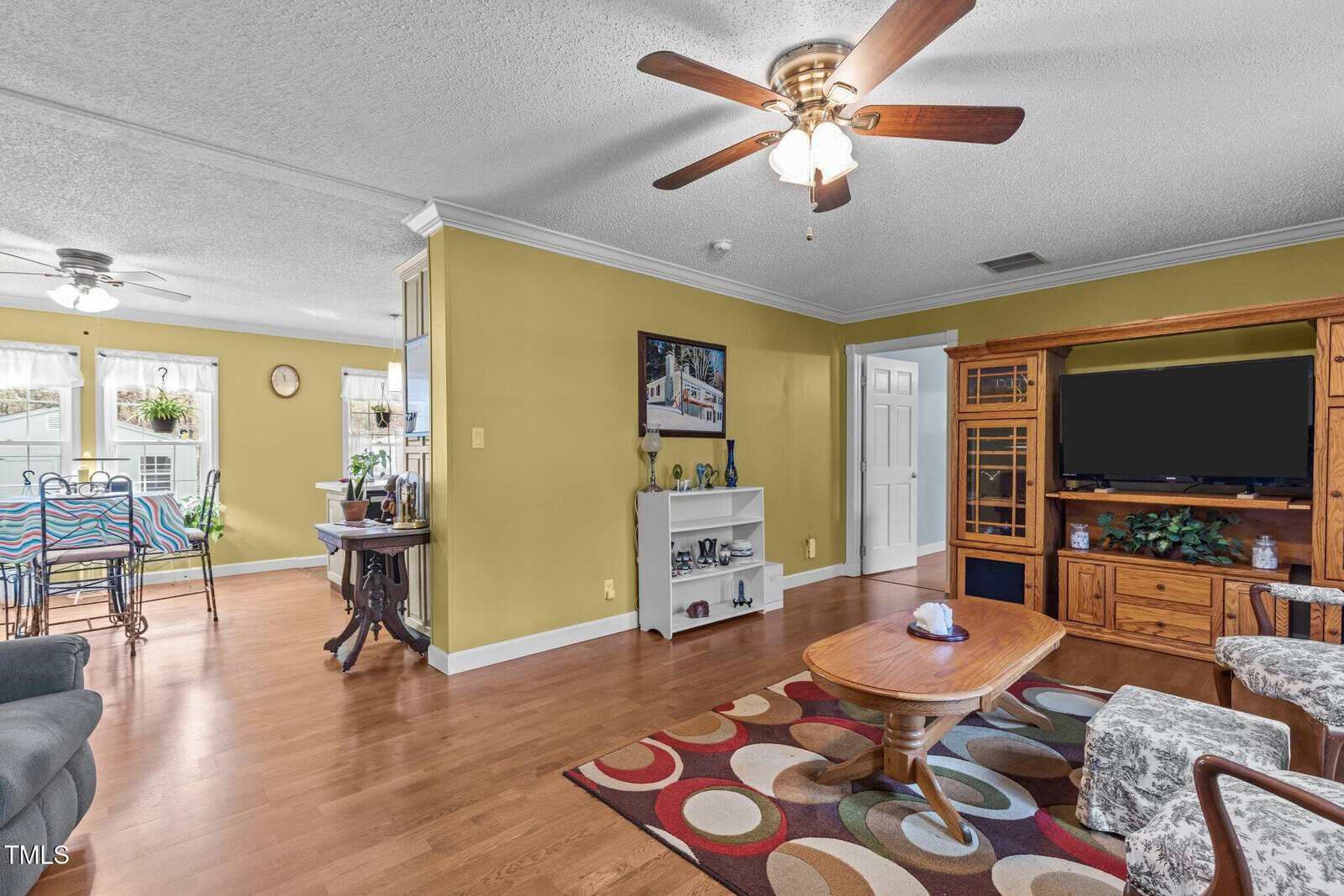 A living room with wood floors, yellow walls, a ceiling fan, and a patterned rug. The room has a wooden TV console, a coffee table, gray upholstered furniture, and an open view into a dining area and kitchen.