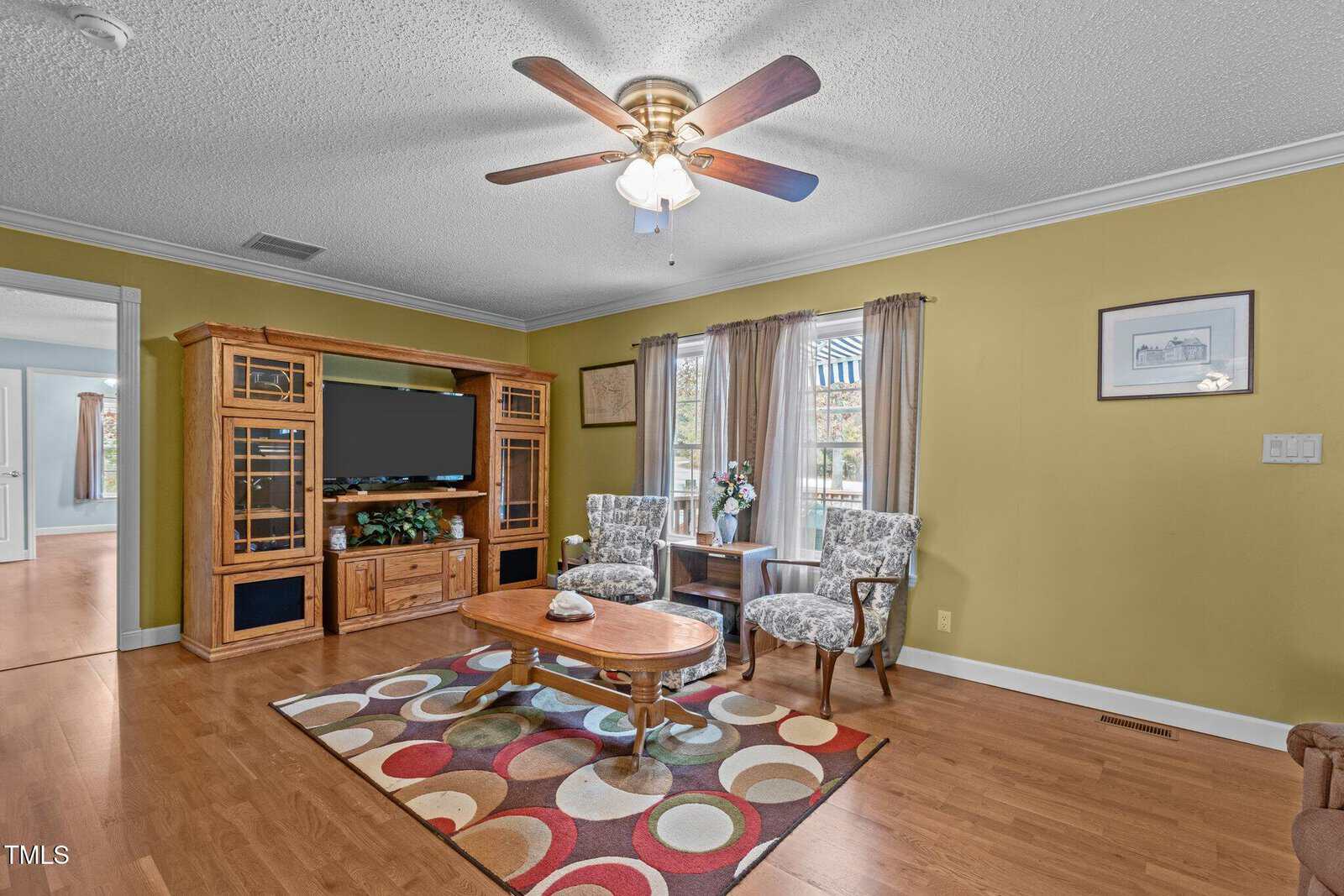 Living room with wood floors, two patterned armchairs, a round table, entertainment center with TV, rug with circles, ceiling fan, and large windows with brown curtains. Walls are painted yellow-green.