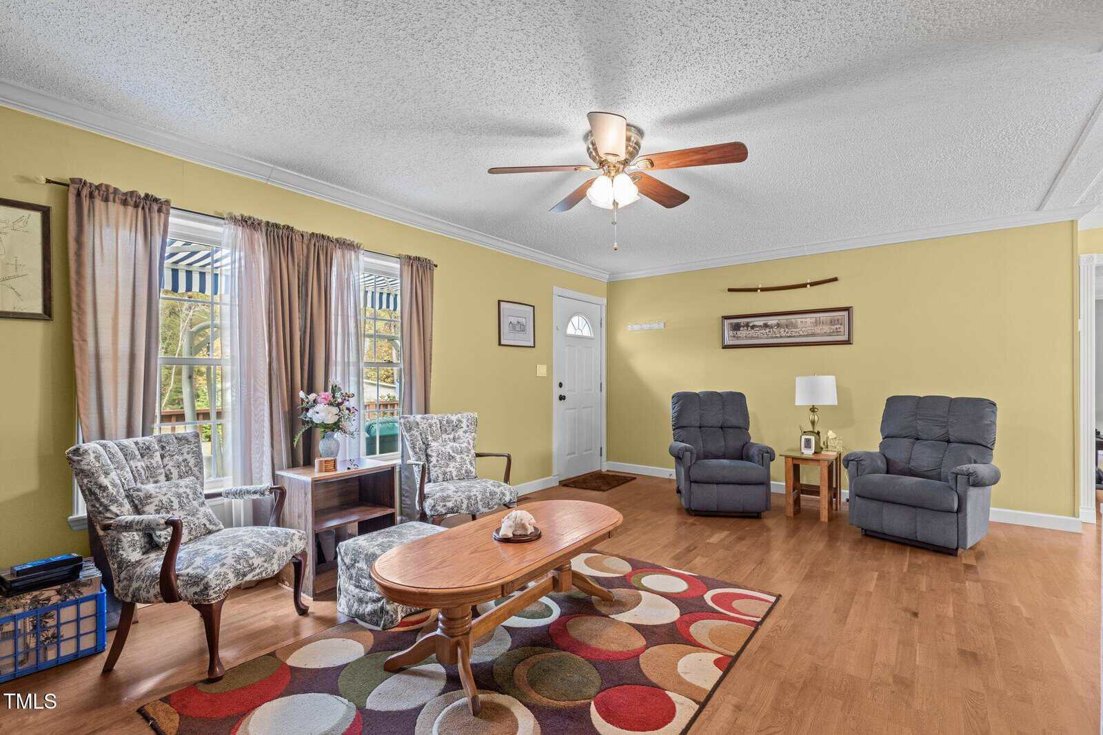 Bright living room with yellow walls, ceiling fan, two armchairs, patterned accent chairs, wooden coffee table, colorful rug, and window with striped awning, allowing natural light into the space.