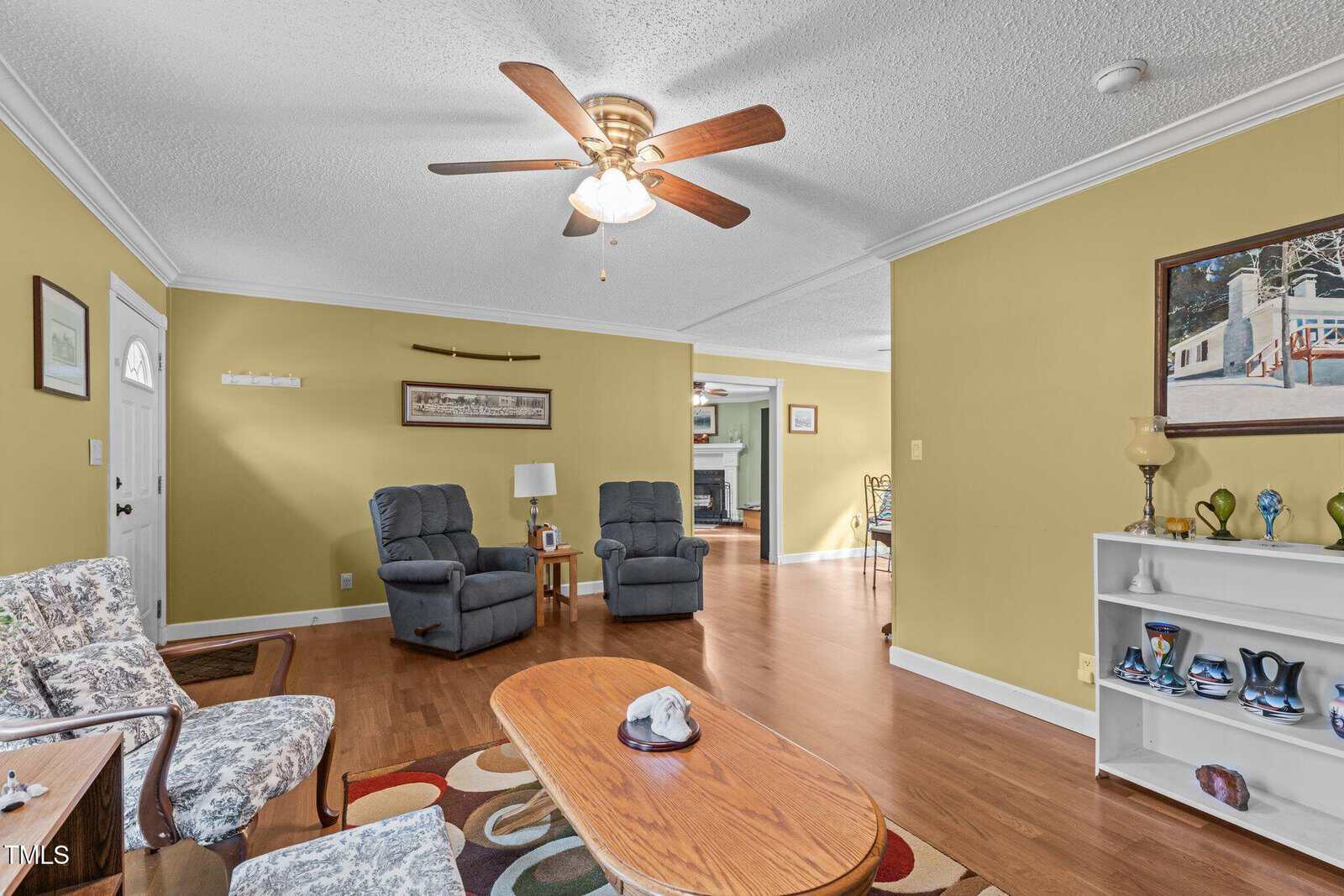 Living room with yellow walls, two armchairs, patterned chairs, a wooden coffee table, a ceiling fan, shelves with decor, and an open view to a dining area and another room in the background.
