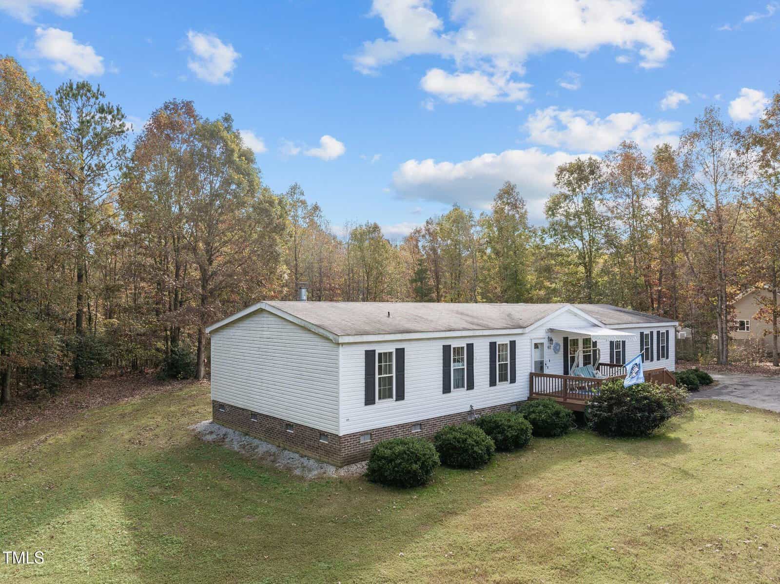 A single-story manufactured home with white siding and brick foundation sits on a grassy lot surrounded by trees. The house has black shutters, a front porch with steps, and a paved driveway. The sky is partly cloudy.