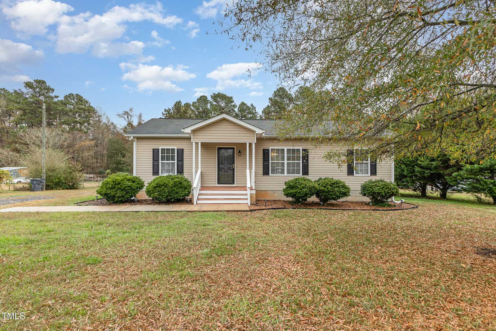 A small single-story beige house with a black roof sits at 4140 Shock Overton Road, surrounded by green shrubbery. A concrete path leads to a central front door with steps. The scene features a grassy lawn and trees in the background under a partly cloudy Oxford sky.