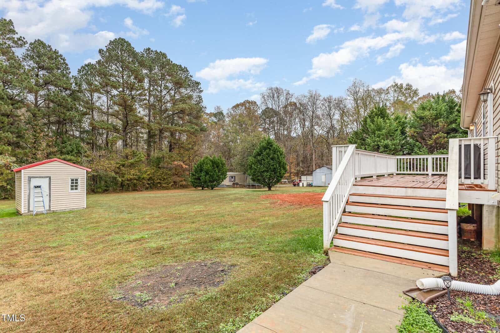 A spacious backyard with a small shed and a wooden deck attached to a house. The yard is grassy with a few trees along the fenced perimeter. A ladder leans against the shed, and the sky is clear with a few clouds.