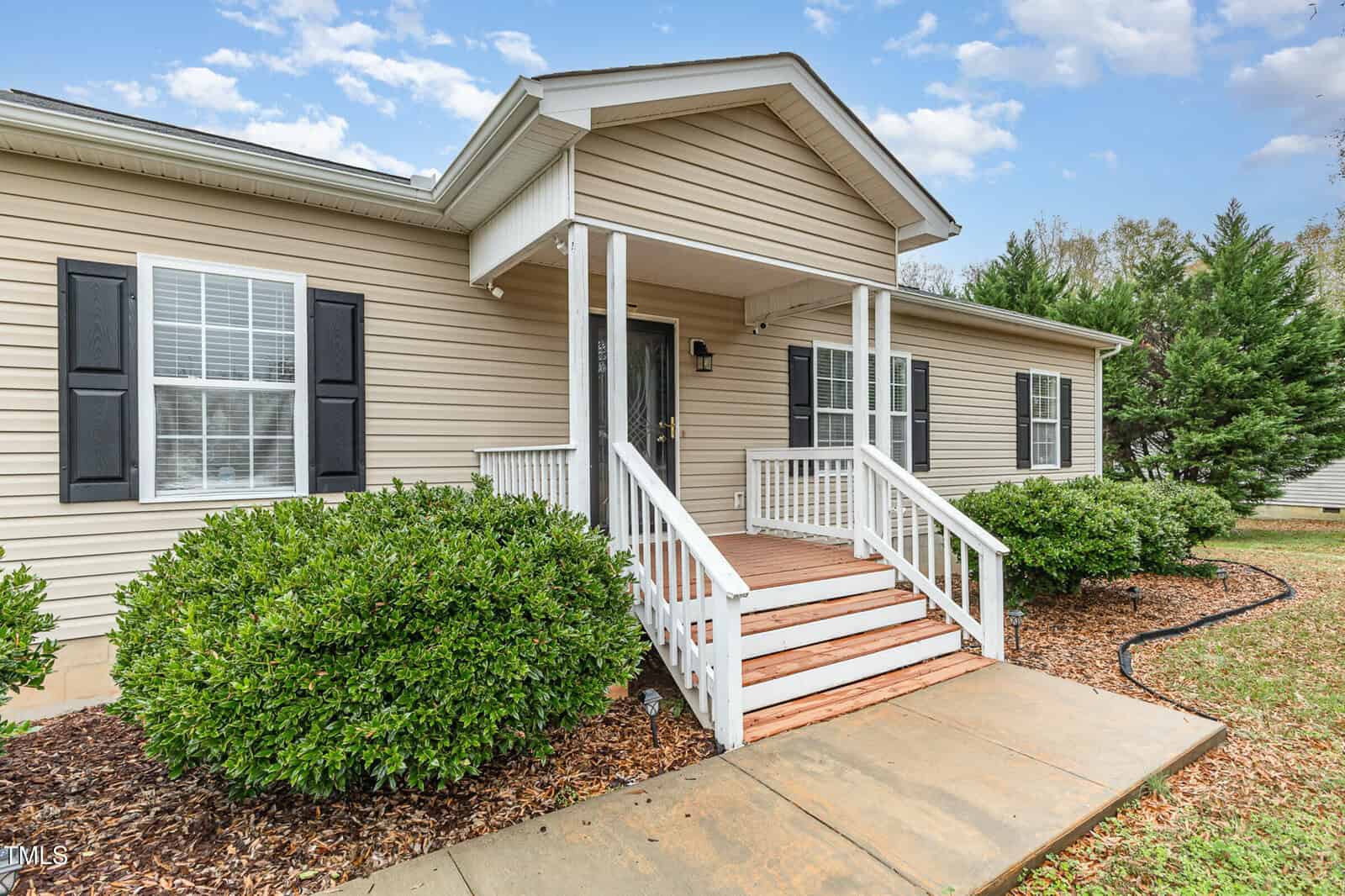 A beige single-story house with a covered front porch, white railings, and steps. It has dark shutters beside the windows. The front yard features green shrubs and a sidewalk leading to the entrance. The sky is partly cloudy.