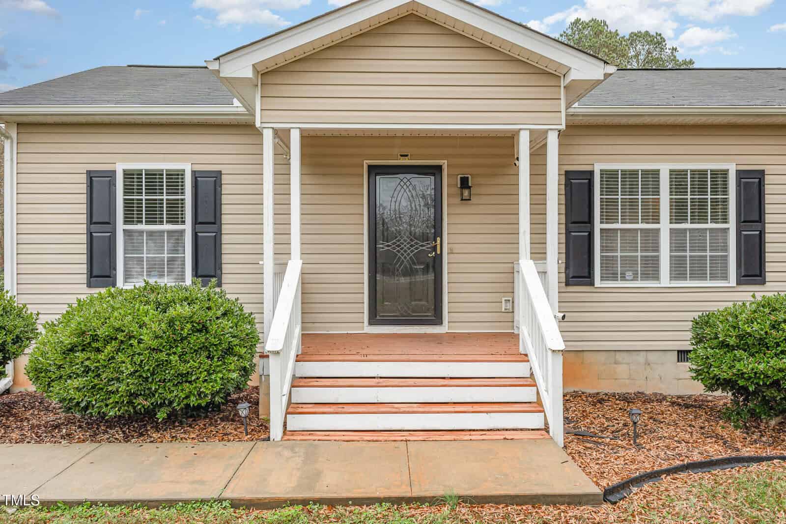 A single-story beige house with a black front door and white trim. The entrance has a small porch with steps and railings. Two windows with black shutters flank the door. Bushes and wood chip landscaping are in front, set against a blue sky.