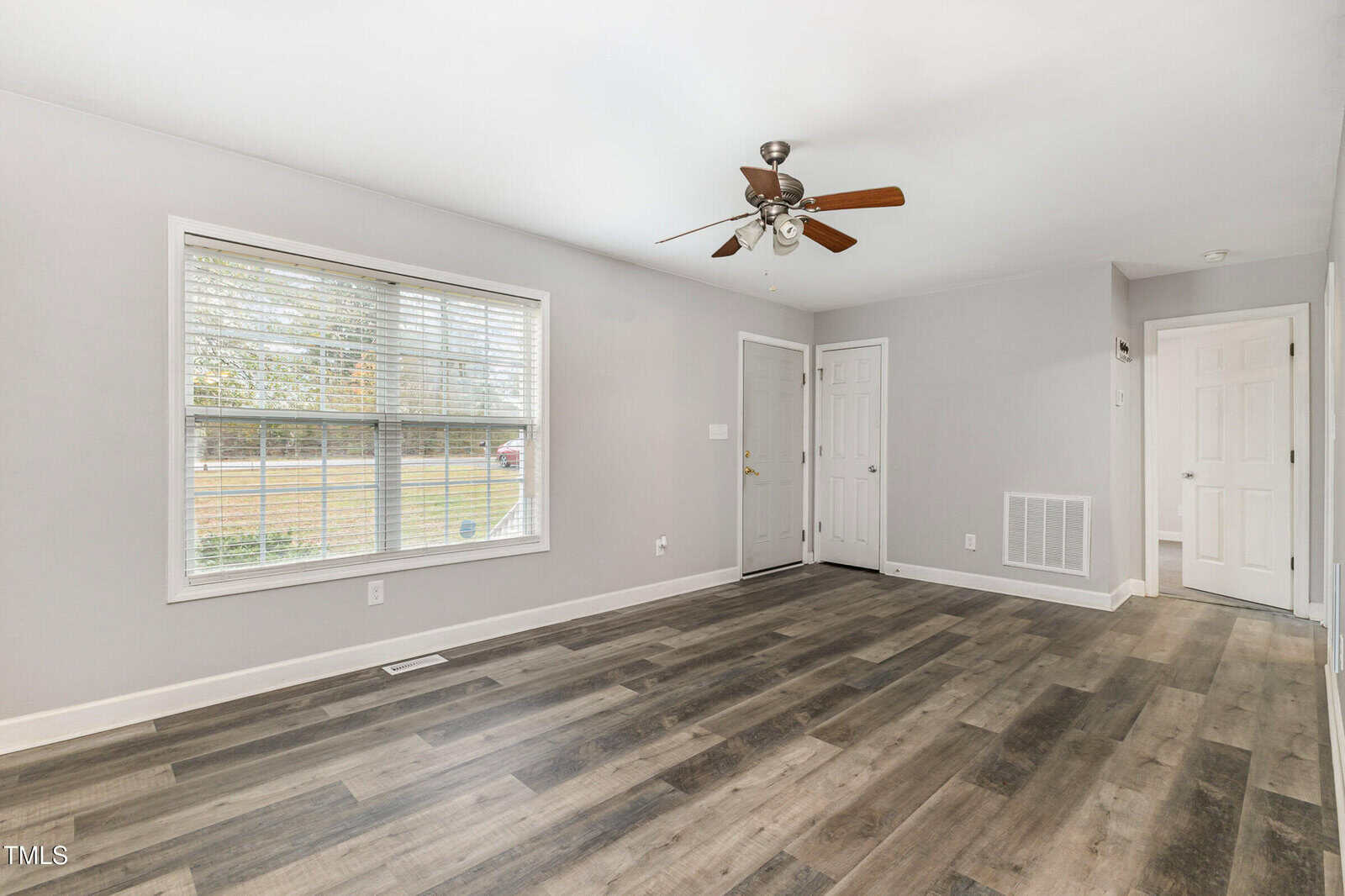 A bright, unfurnished room with gray walls and wood-look vinyl flooring. It features a large window with blinds, a ceiling fan, and multiple doors. The room is well-lit and spacious.