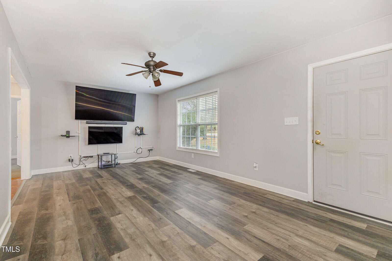 A spacious living room with light gray walls and dark wood flooring. It features a ceiling fan, a wall-mounted TV, and a window with white blinds. A door is on the right, and an adjoining room is visible through a doorway on the left.