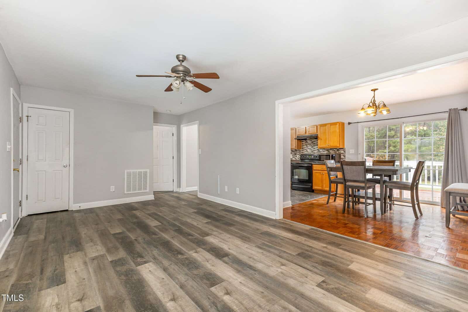 Spacious open-concept living area with light gray walls, wood-like flooring, and a ceiling fan. The adjacent dining area features wooden furniture and large windows overlooking a lush green outdoor view.