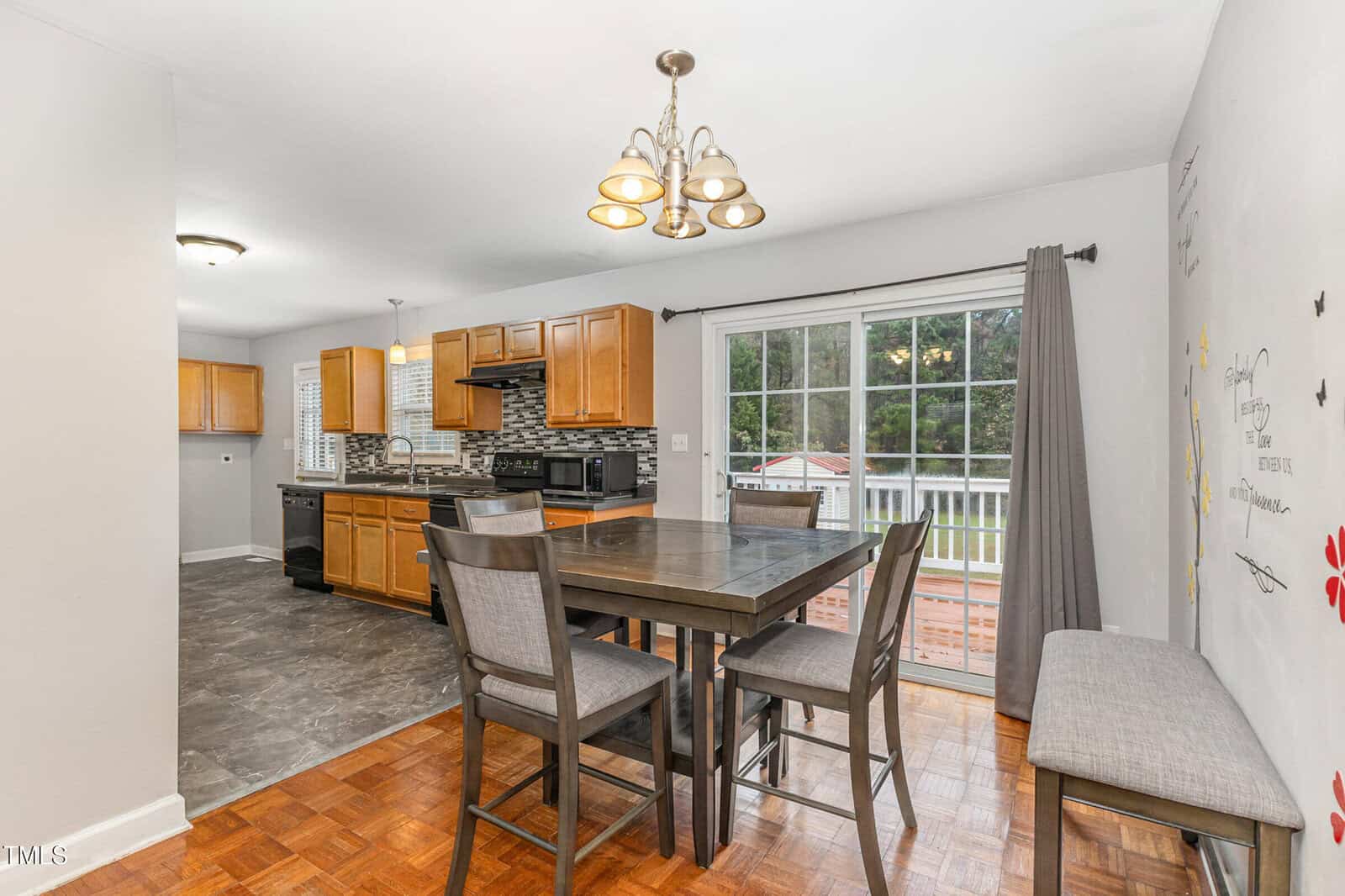 Kitchen and dining area with wooden cabinets, a tiled backsplash, and a central island. There's a dark dining table with four chairs, large glass doors leading to an outside view, and a chandelier hanging from the ceiling.