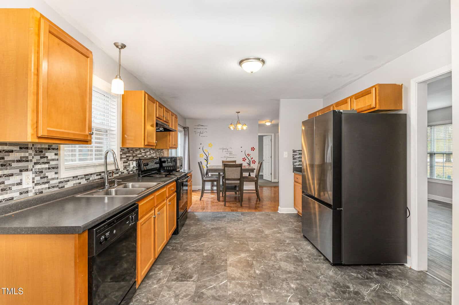 A modern kitchen with wooden cabinets, black appliances, and a mosaic tile backsplash. A dining area with a table and chairs is visible in the background. The floor is tiled in a gray pattern, and the walls are painted white.