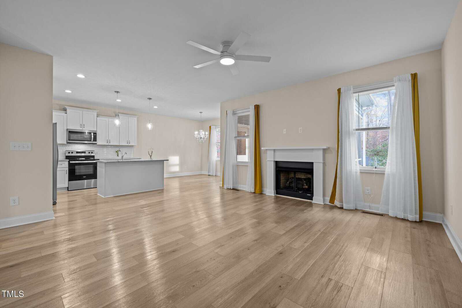 Open-concept living room and kitchen with light wood floors, white cabinets, a ceiling fan, fireplace, and large windows with white curtains and yellow accents. Modern and bright interior.