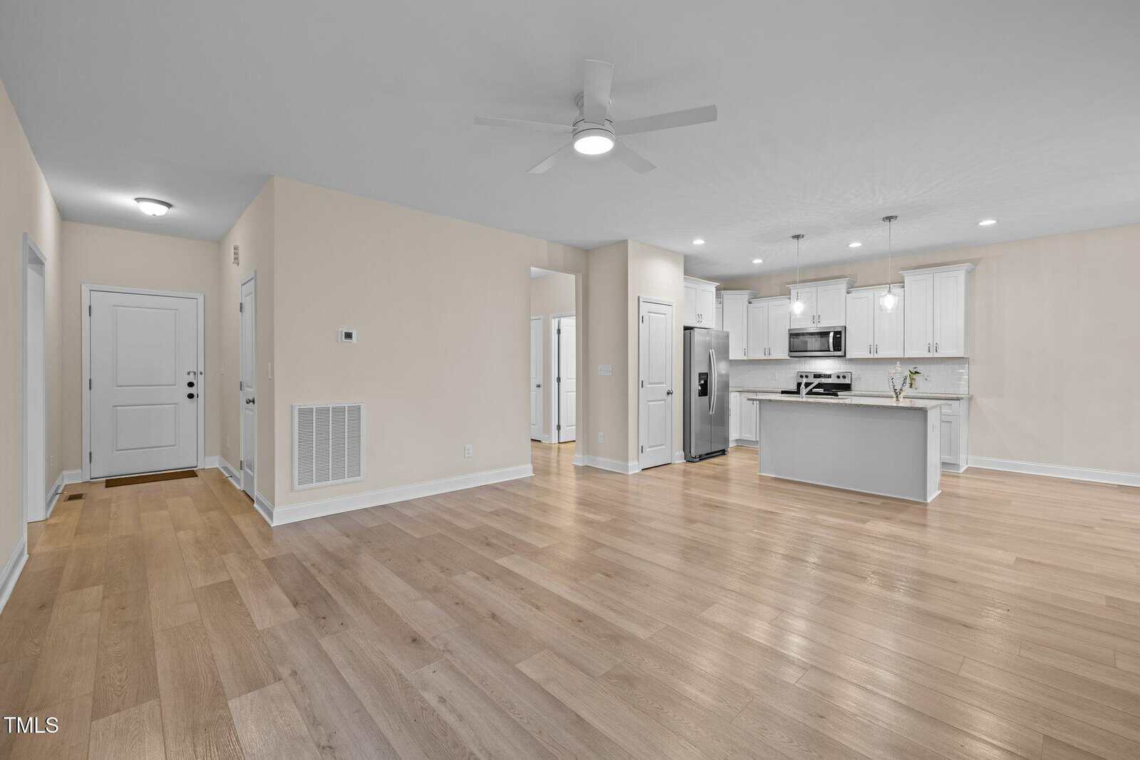 Open modern living space with light wood floors, beige walls, white trim, ceiling fan, and view of a kitchen with white cabinets, stainless steel appliances, and an island with pendant lights.