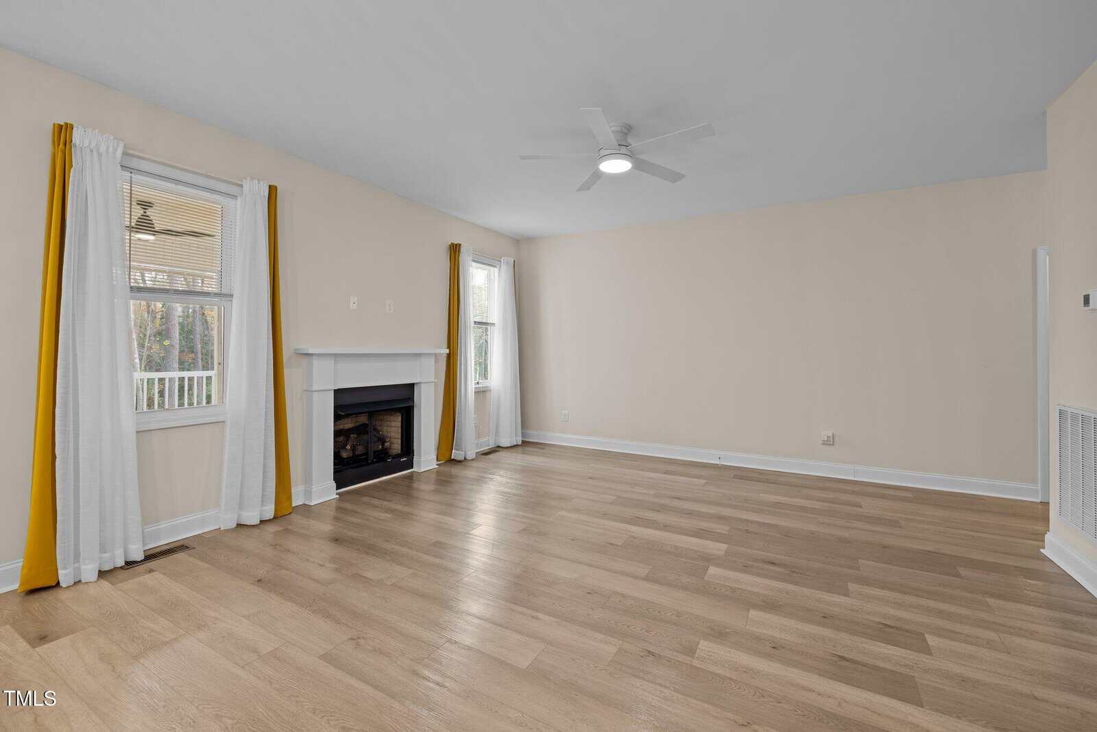 Spacious living room with light wood floors, beige walls, a white ceiling fan, two windows with white and yellow curtains, and a white fireplace with a black insert.