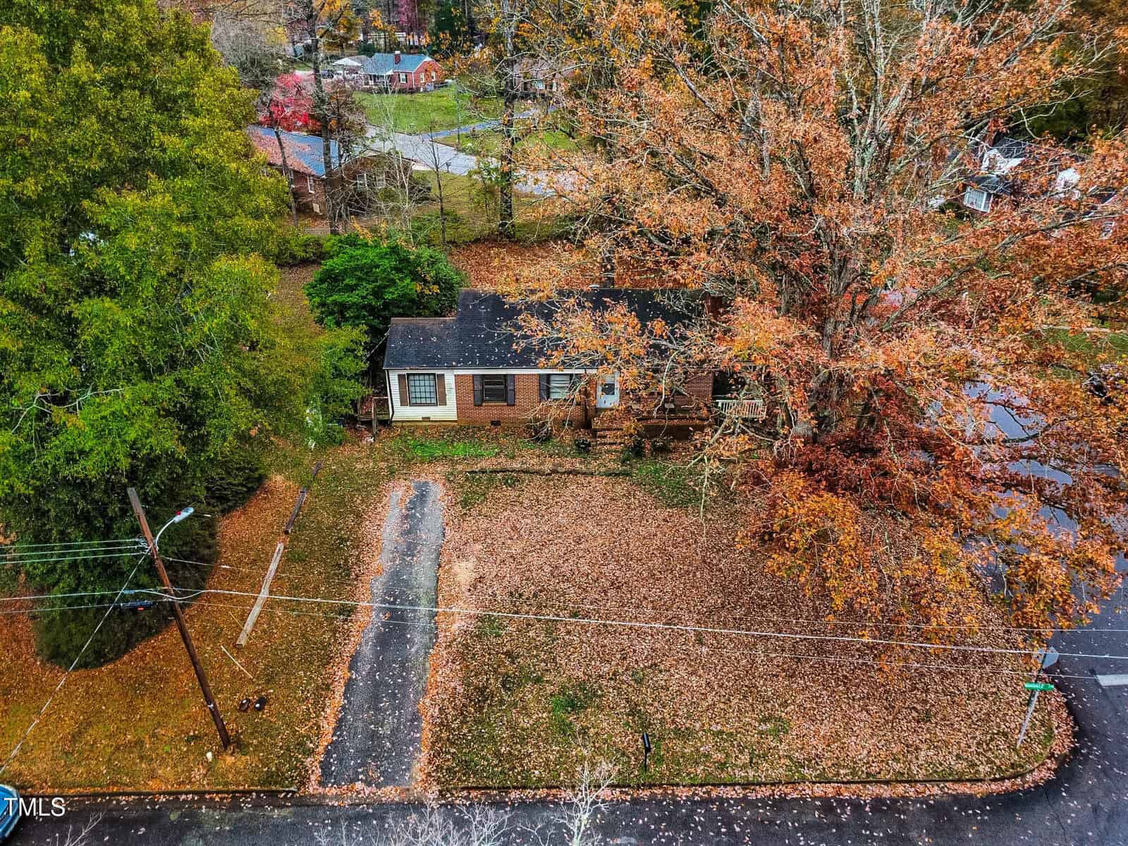 Aerial view of a brick house surrounded by autumn trees. The yard is covered in fallen leaves, and a driveway leads to the house. Nearby, other homes and streets can be seen amid the colorful foliage.