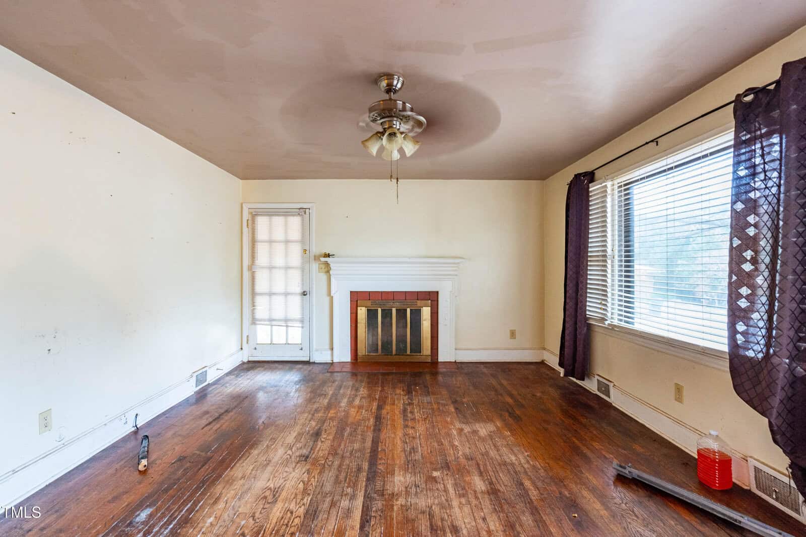 A vacant living room with hardwood floors and a ceiling fan. There are cream-colored walls, a white fireplace with a gold-rimmed firewood cover, and a red bucket in the corner. Purple curtains partially cover the large window on the right.