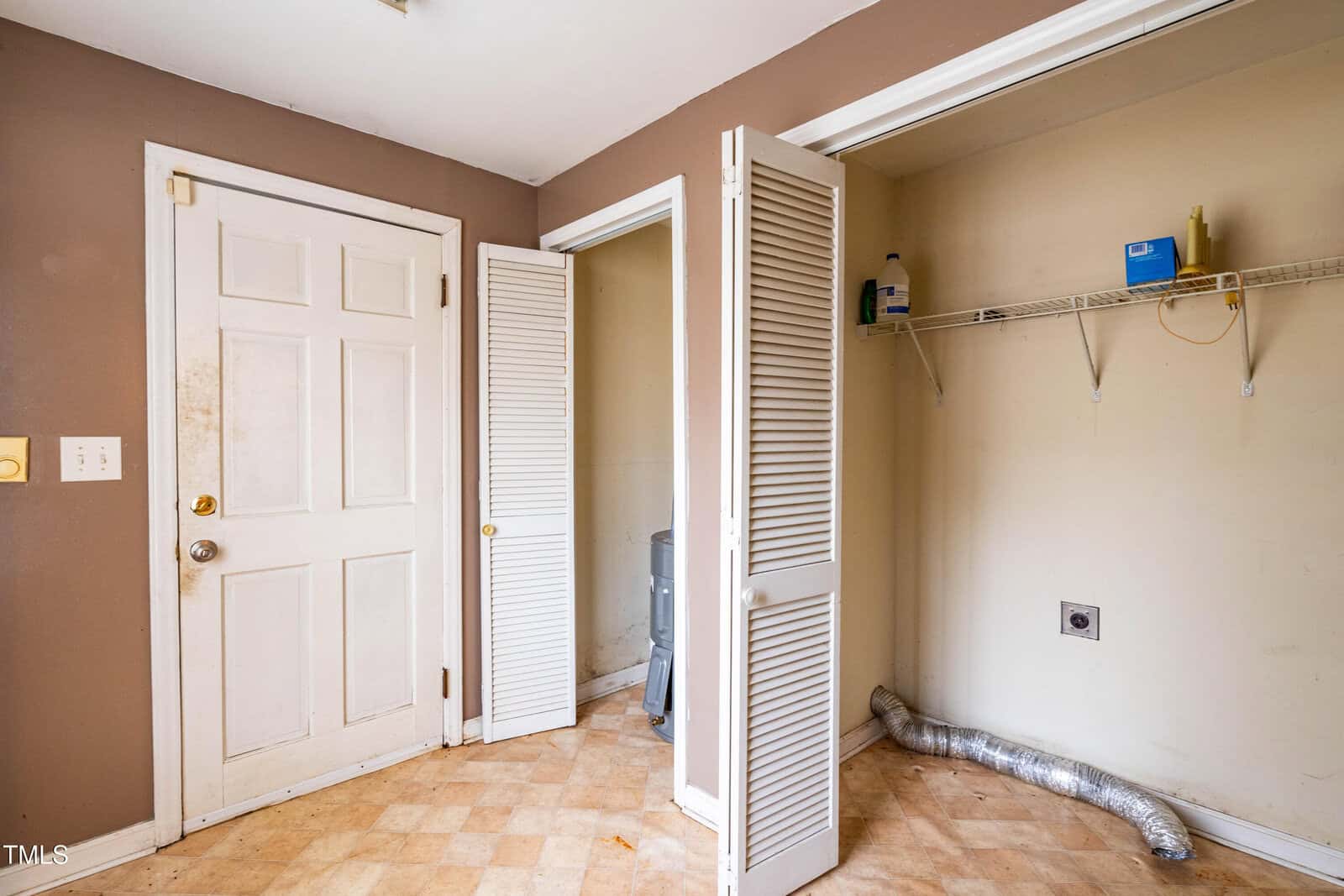 Empty laundry room with brown walls, a closed white door, folding closet doors, a wire shelf with cleaning supplies, and a dryer vent hose on the floor. The floor has a light checkered pattern.