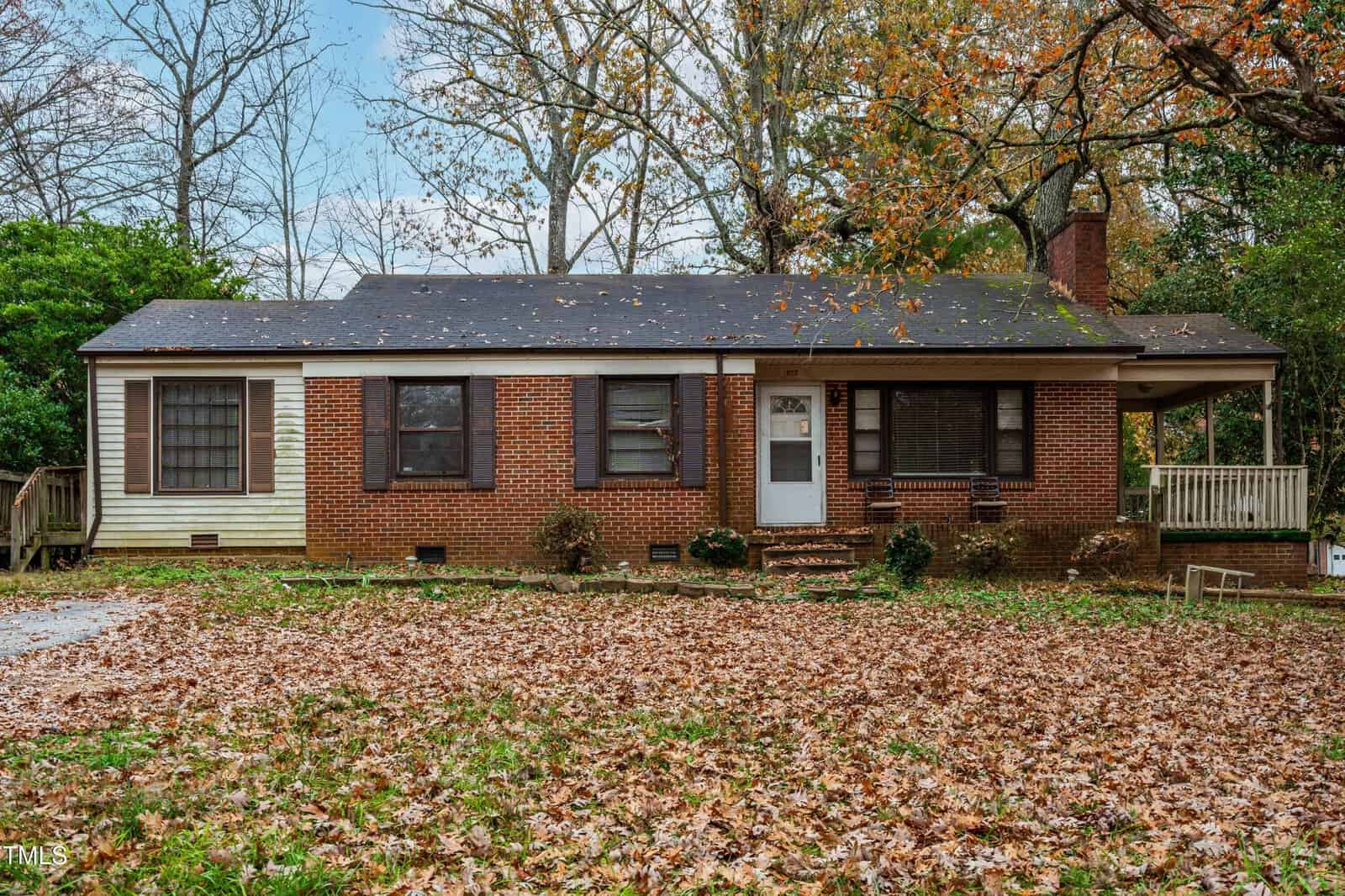Nestled at 1655 Oakdale Circle in Henderson, this charming single-story red brick house features a dark roof and brown shutters. The yard, blanketed with autumn leaves, is dotted with trees, and a welcoming white door graces the front porch steps.
