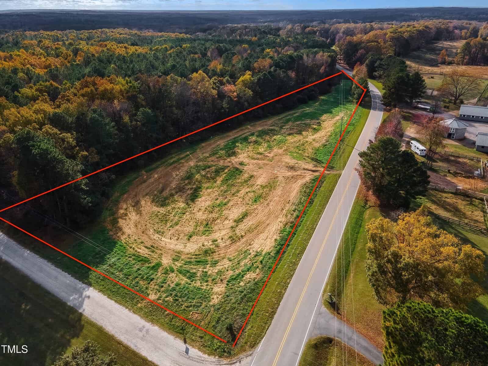 Aerial view of a triangular plot of land bordered by red lines. The area is mostly cleared with surrounding trees. Two roads run along its edges, and there are nearby buildings. The landscape is captured during autumn with colorful foliage.