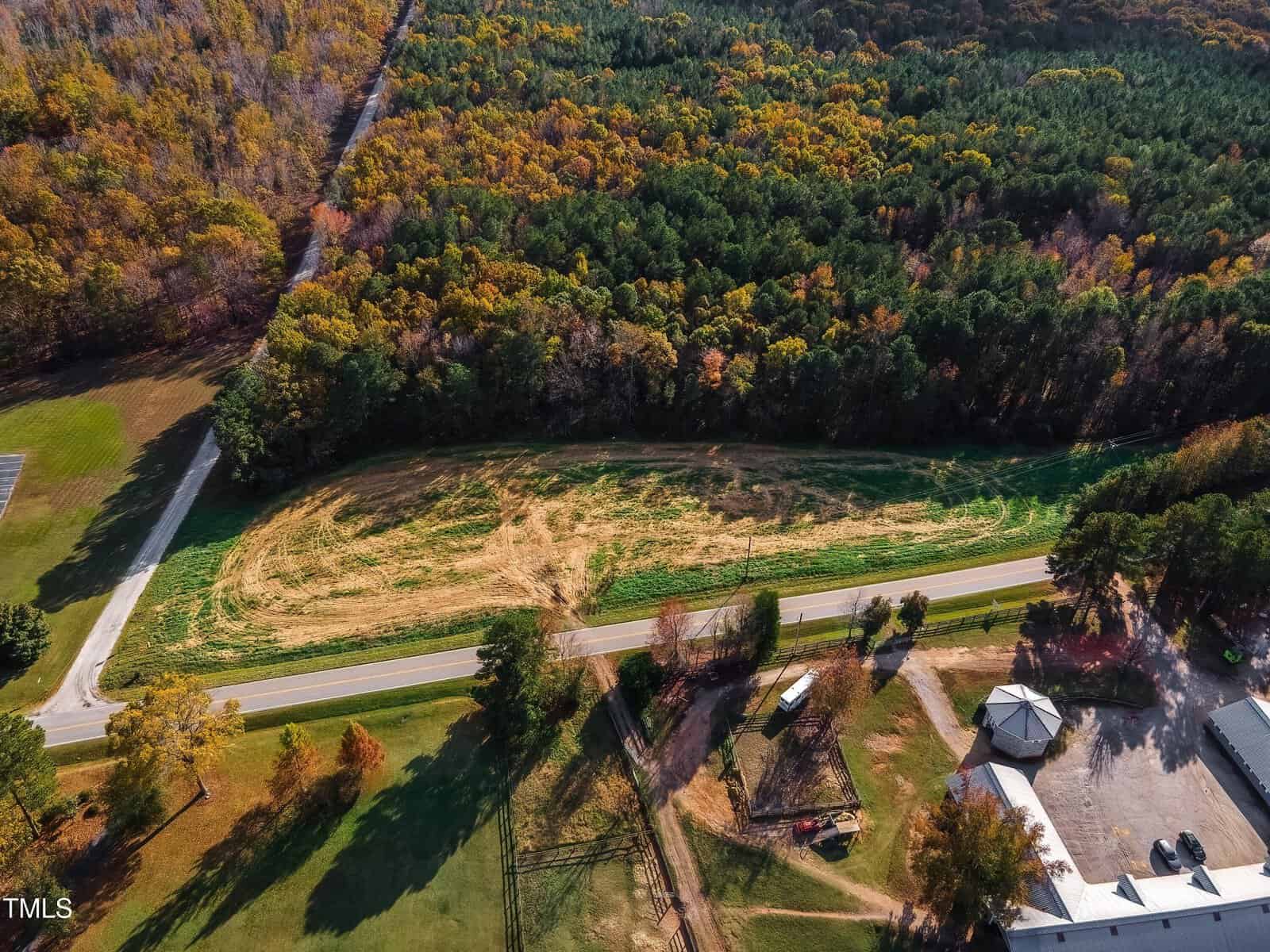 Aerial view of a curving two-lane road bordered by trees and a grassy field with some cleared patches. Buildings and a fenced area are visible in the foreground. Dense forest surrounds the scene.