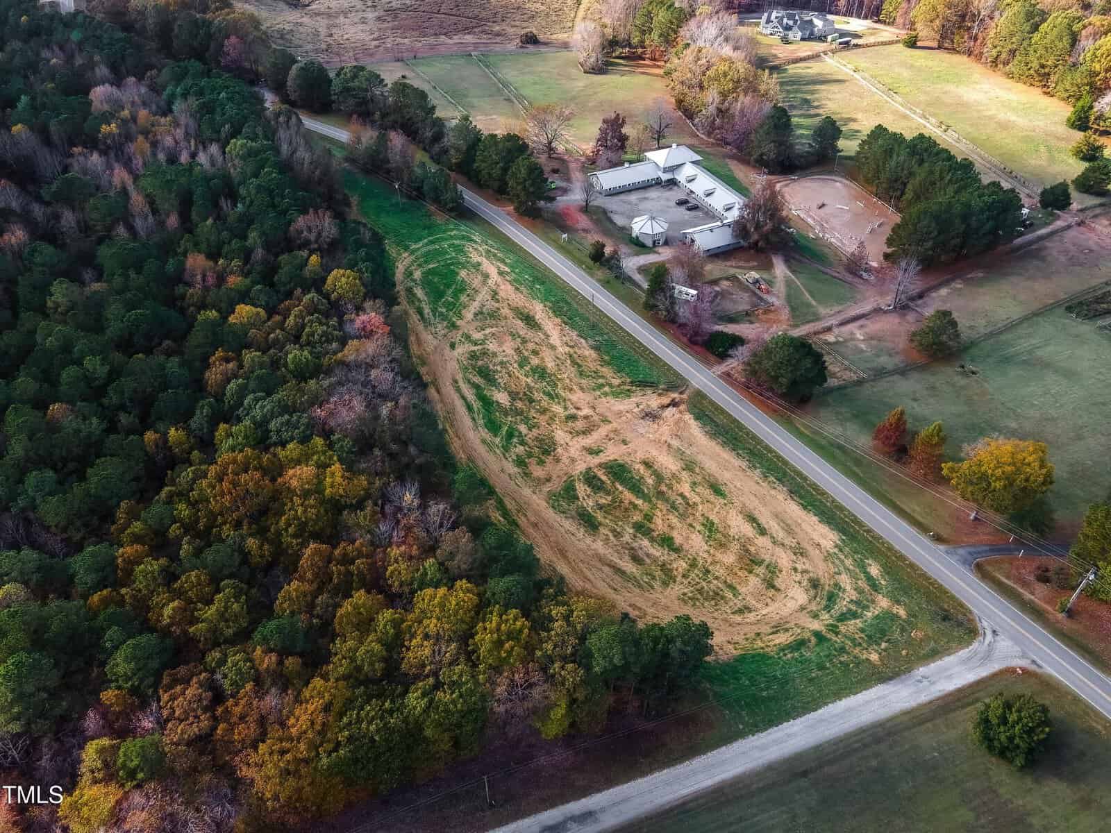 Aerial view of a landscape featuring a grassy field with a zigzag path, surrounded by dense woods. Nearby, a road runs parallel to the field, and a building complex with multiple structures is visible in the upper right corner.