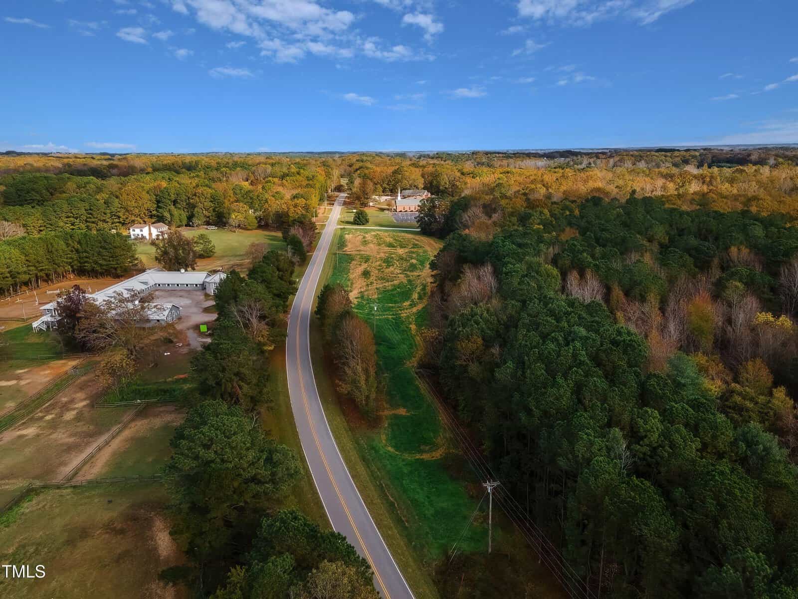 Aerial view of a rural landscape with a winding road stretching through a dense forest and fields. A few buildings are visible on the left, surrounded by greenery. The sky is clear with scattered clouds.