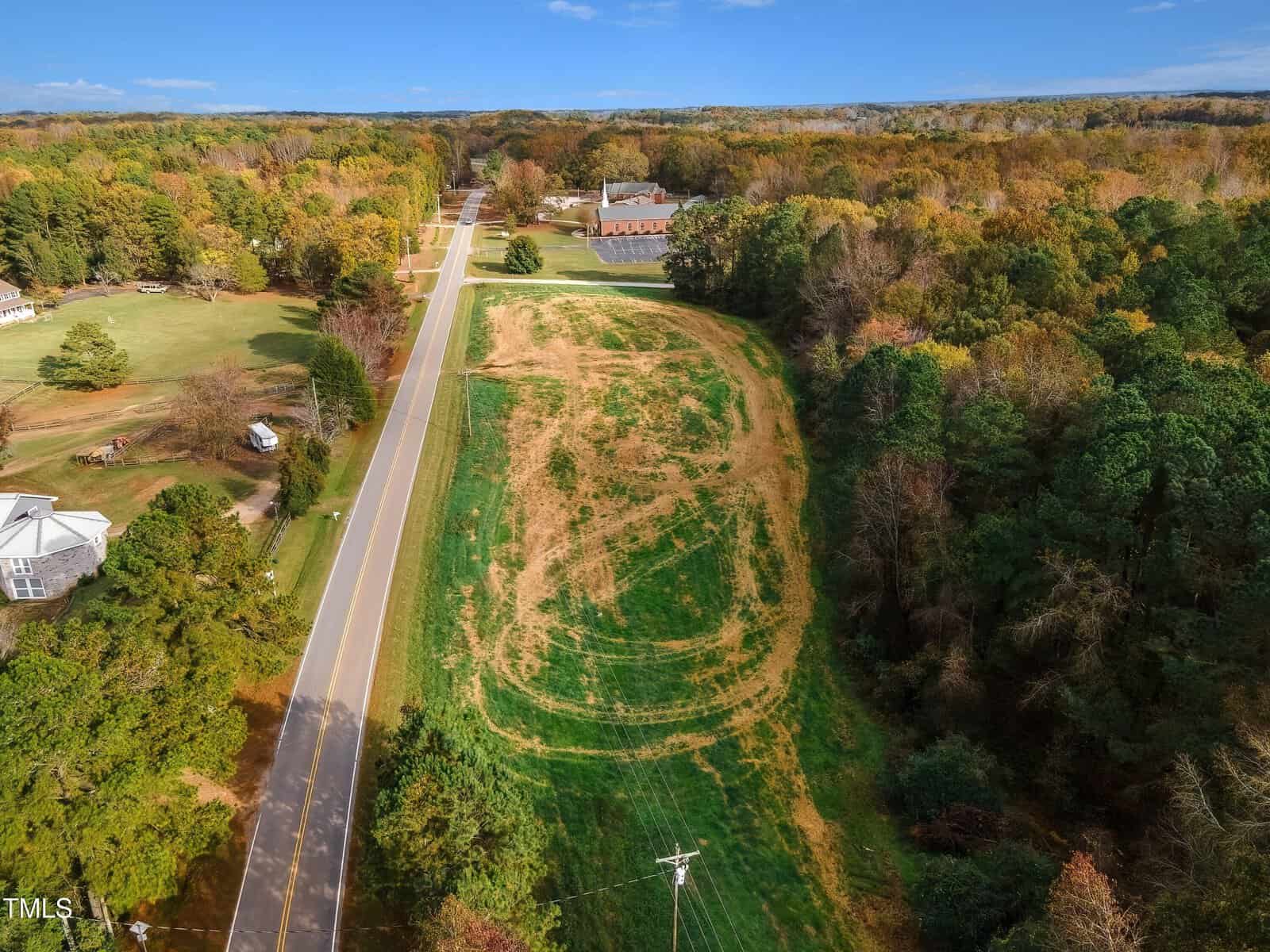 Aerial view of a rural landscape featuring a road lined with trees on one side and an open field on the other. There are a few scattered buildings, including a large one in the distance. The scene is surrounded by forested areas and hills.