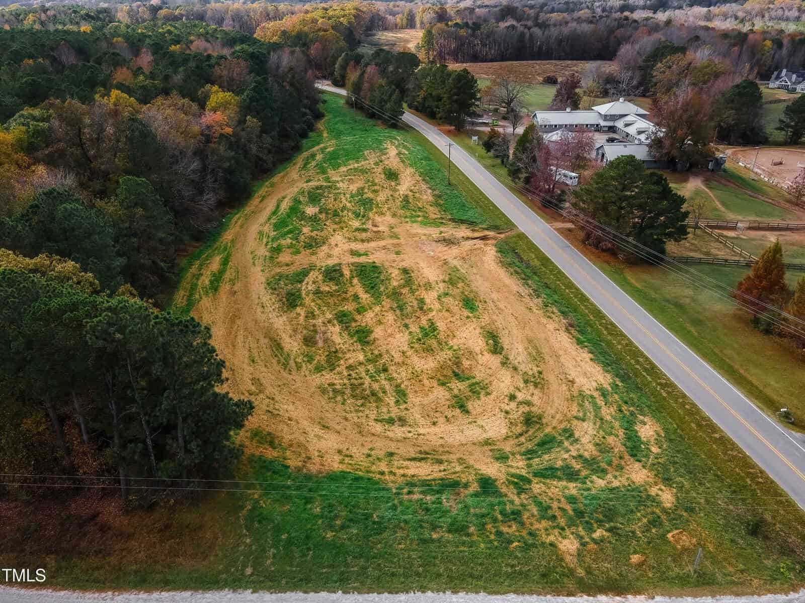 An aerial view of a grassy field with a circular patch of sparse vegetation. A road runs parallel to the field, bordered by trees on one side and buildings in the background. The scene is surrounded by a mix of autumn-colored and evergreen trees.