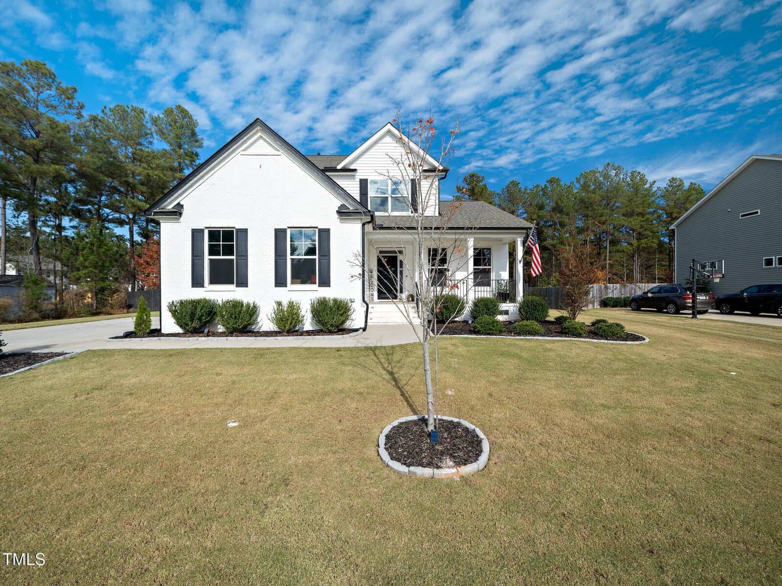 A white two-story suburban house with black shutters and a covered porch, set on a large lawn with trimmed bushes and a small tree in the front yard. The sky is partly cloudy and other houses are visible nearby.