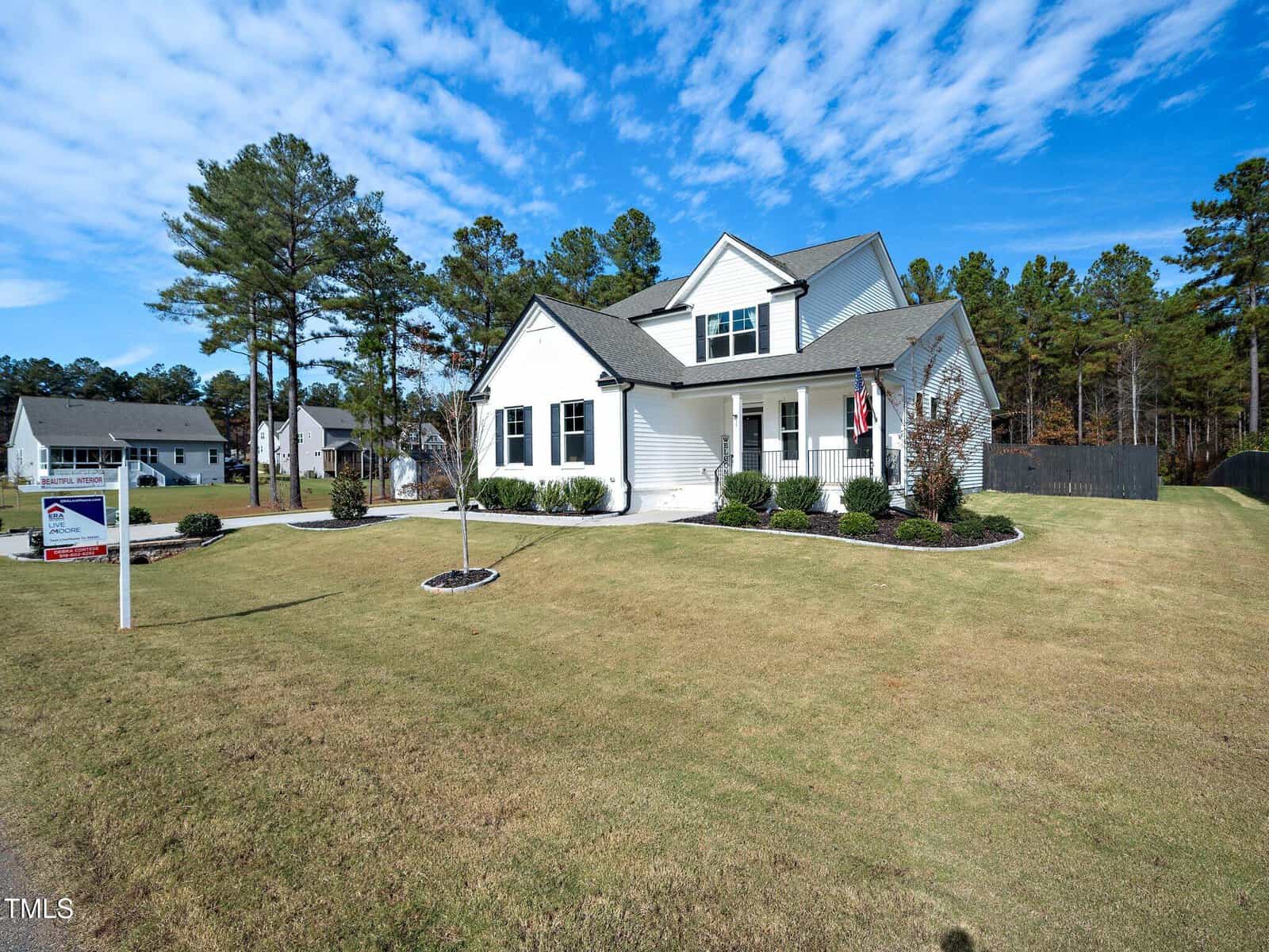 A white two-story house with a large, well-kept lawn, surrounded by trees and a blue sky with scattered clouds. A “for sale” sign is posted near the front of the property.