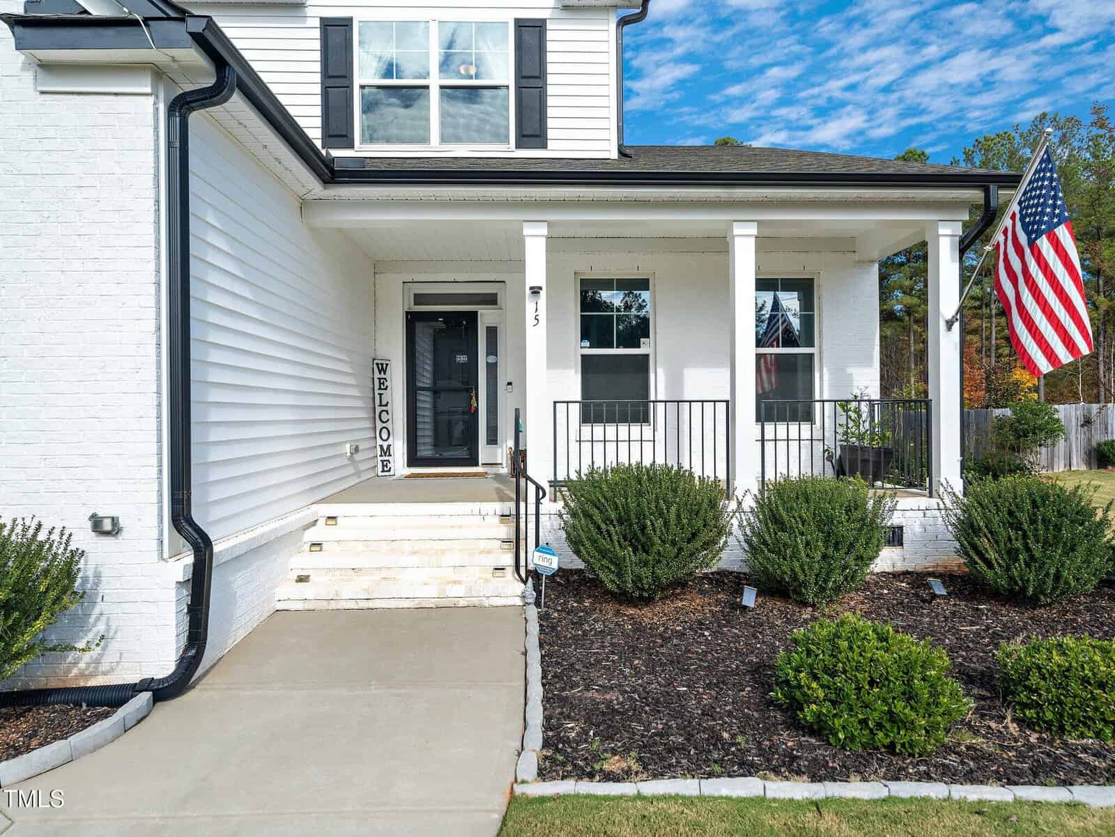 A white house with black shutters and trim features a covered front porch, four white columns, a "Welcome" sign by the door, an American flag, trimmed bushes, and a concrete walkway leading to the steps.
